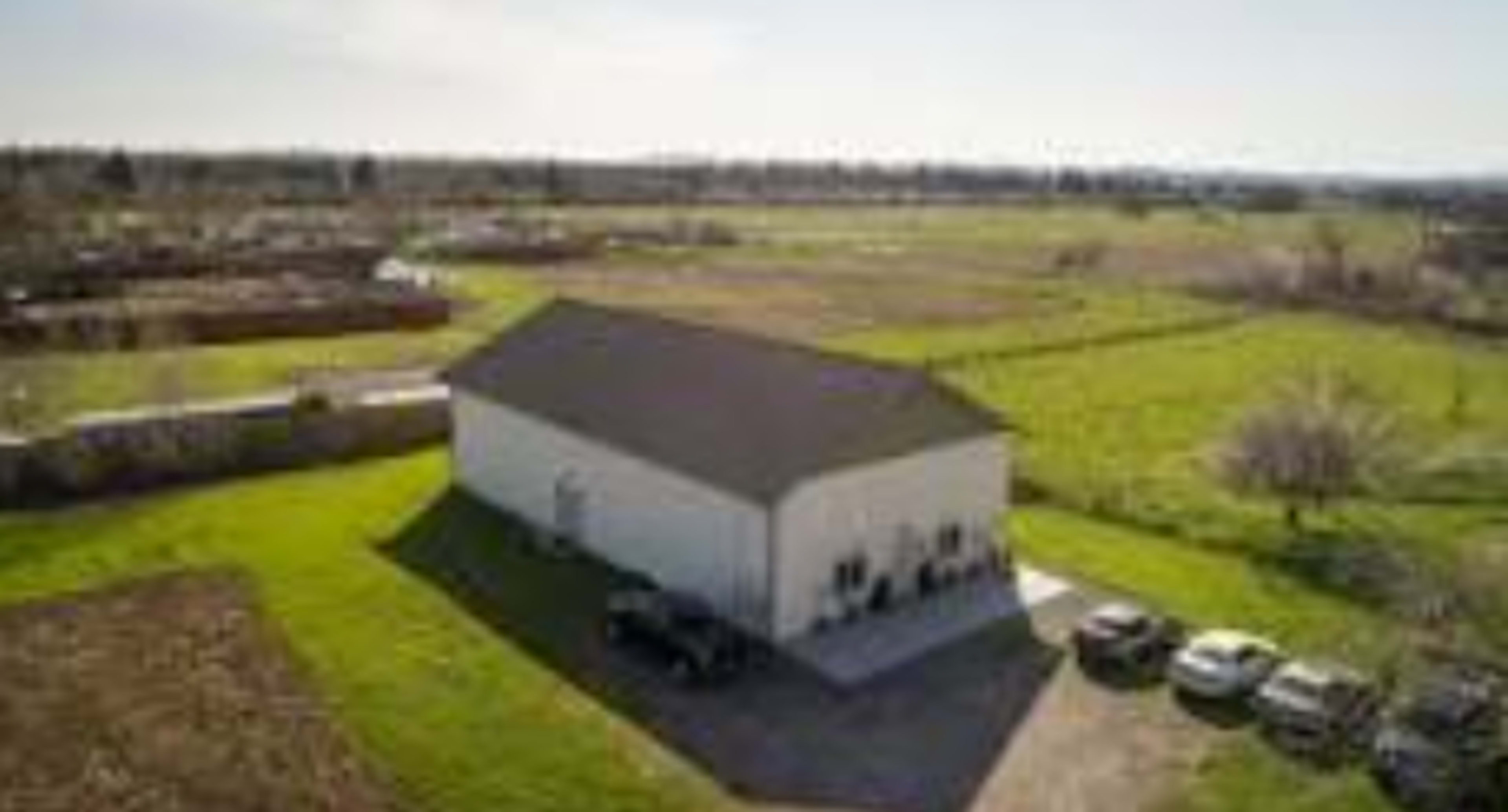A large white building sits on a green expanse surrounded by rural farmland and parked vehicles.