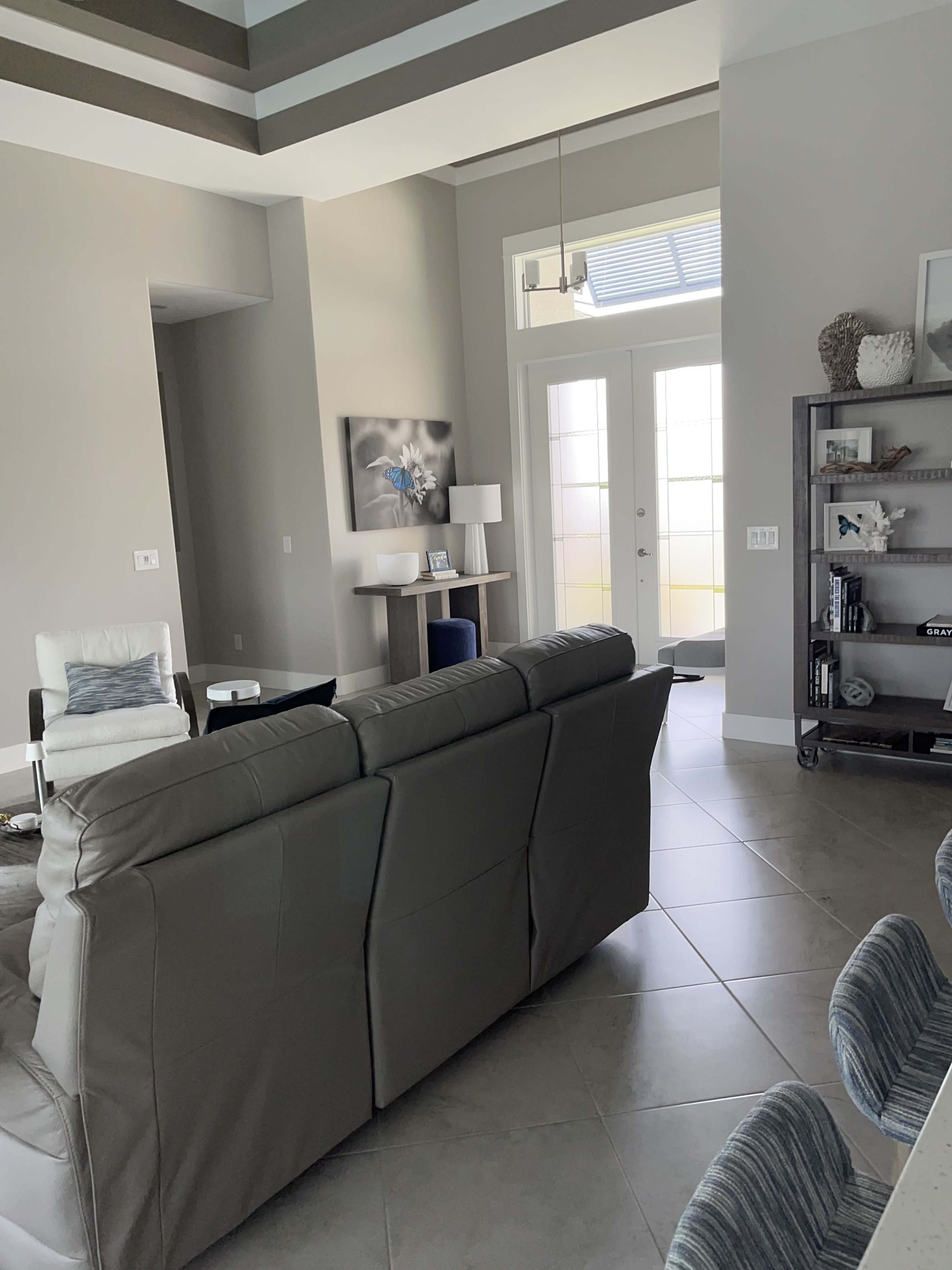 A modern living room features a beige leather sofa facing an entrance with large glass doors, surrounded by light-colored walls and minimalistic furnishings.