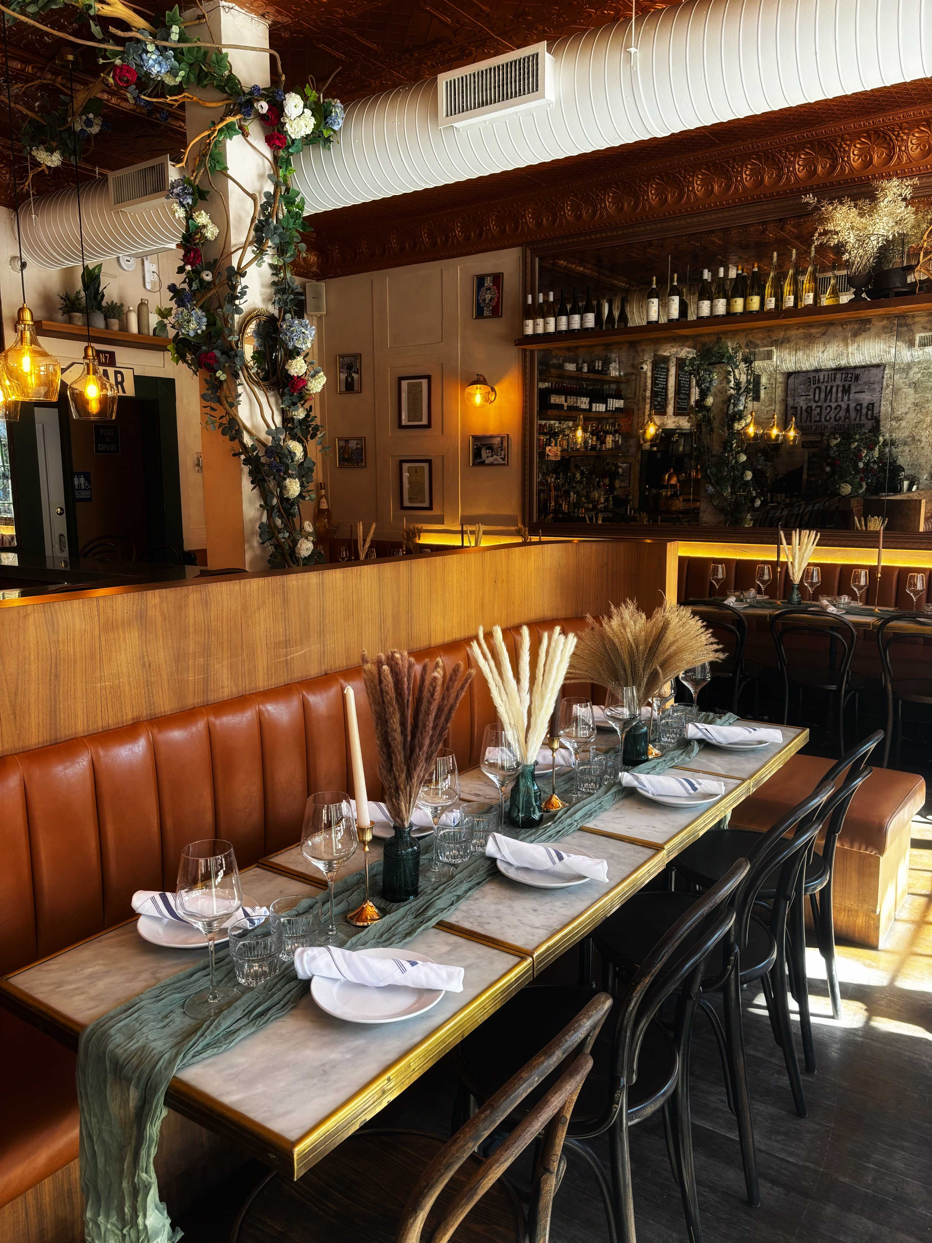 A long dining table is set with elegant tableware and centerpieces made of dried plants, surrounded by a stylish restaurant interior featuring exposed brick and decorative shelving.