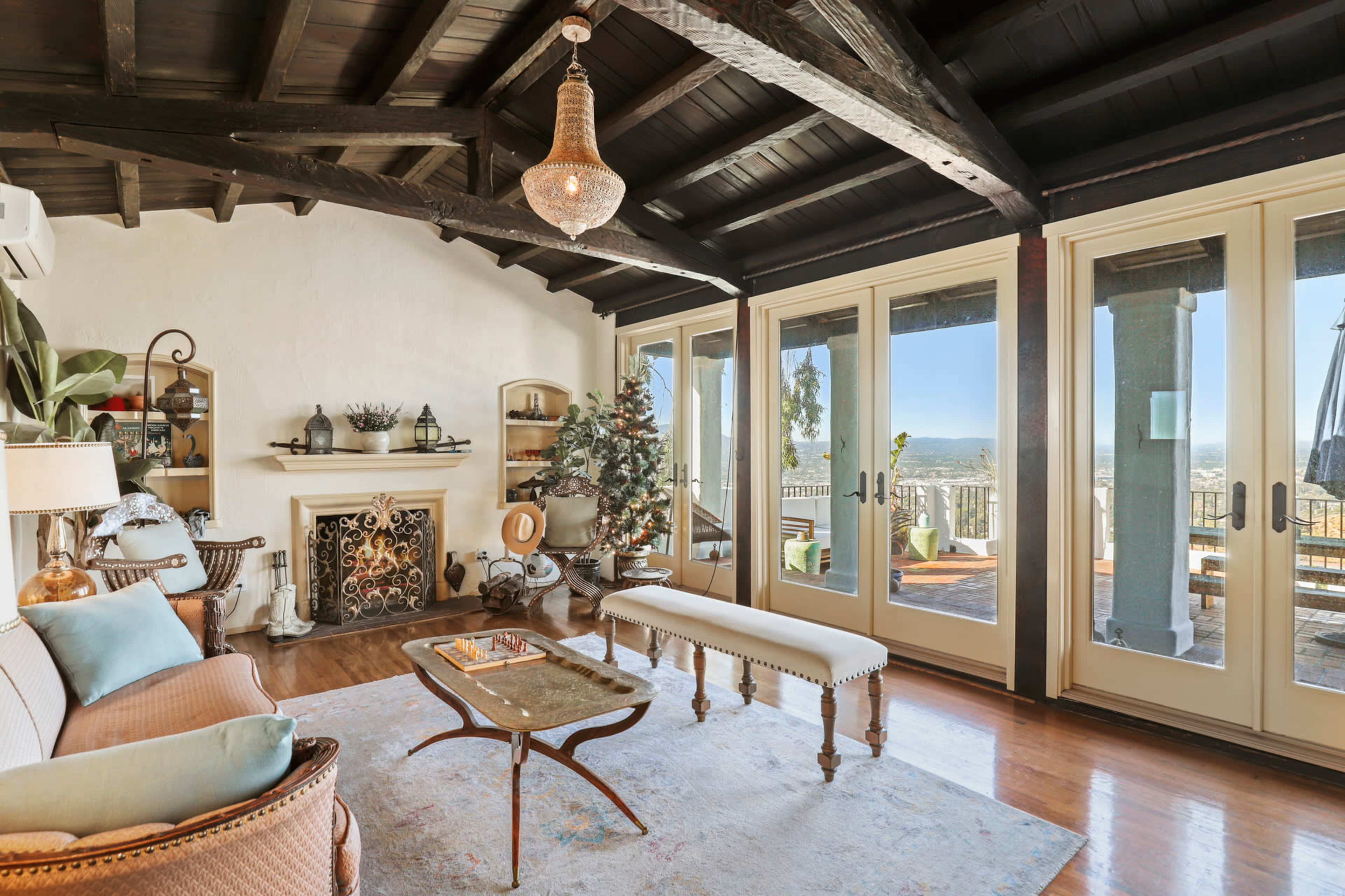A cozy living room features a wooden ceiling, large glass doors leading to a balcony, and a decorative fireplace surrounded by furniture and a Christmas tree.