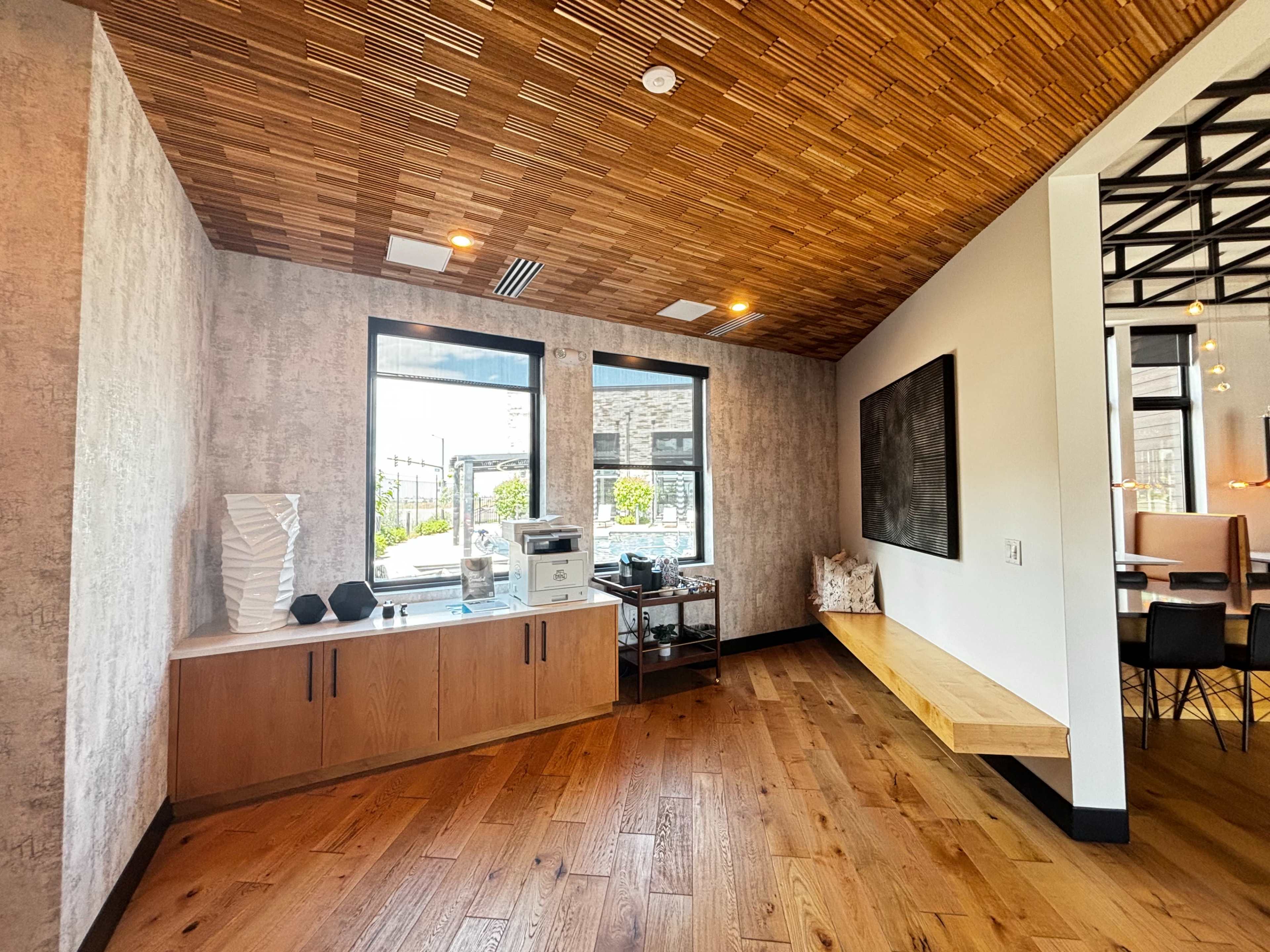 A modern interior space with a wooden ceiling, large windows, and a minimalistic design featuring a wooden bench and a countertop area.