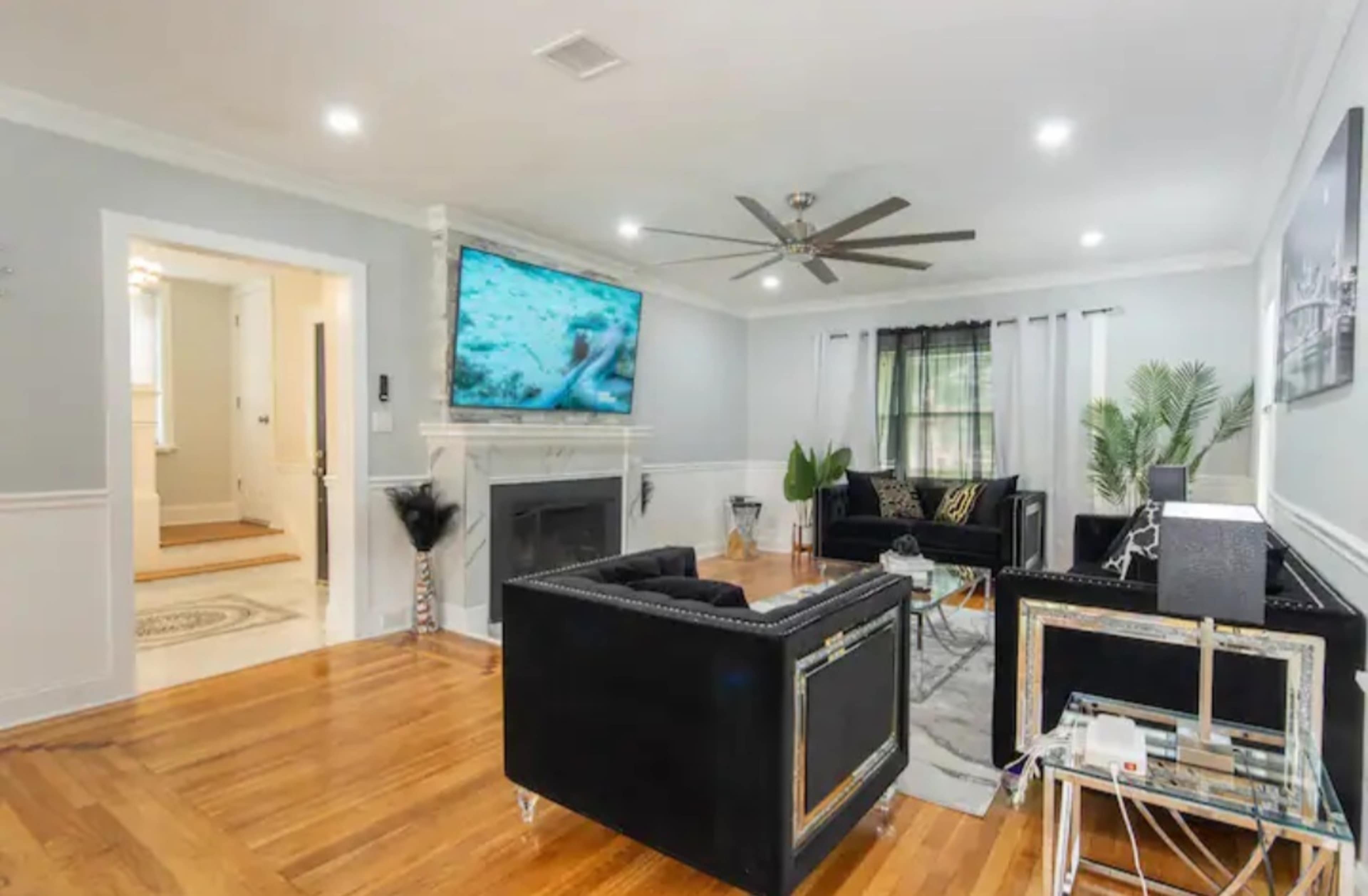 The living room features a black leather sofa set, a wall-mounted television, and hardwood floors, with a doorway leading to a hallway in the background.