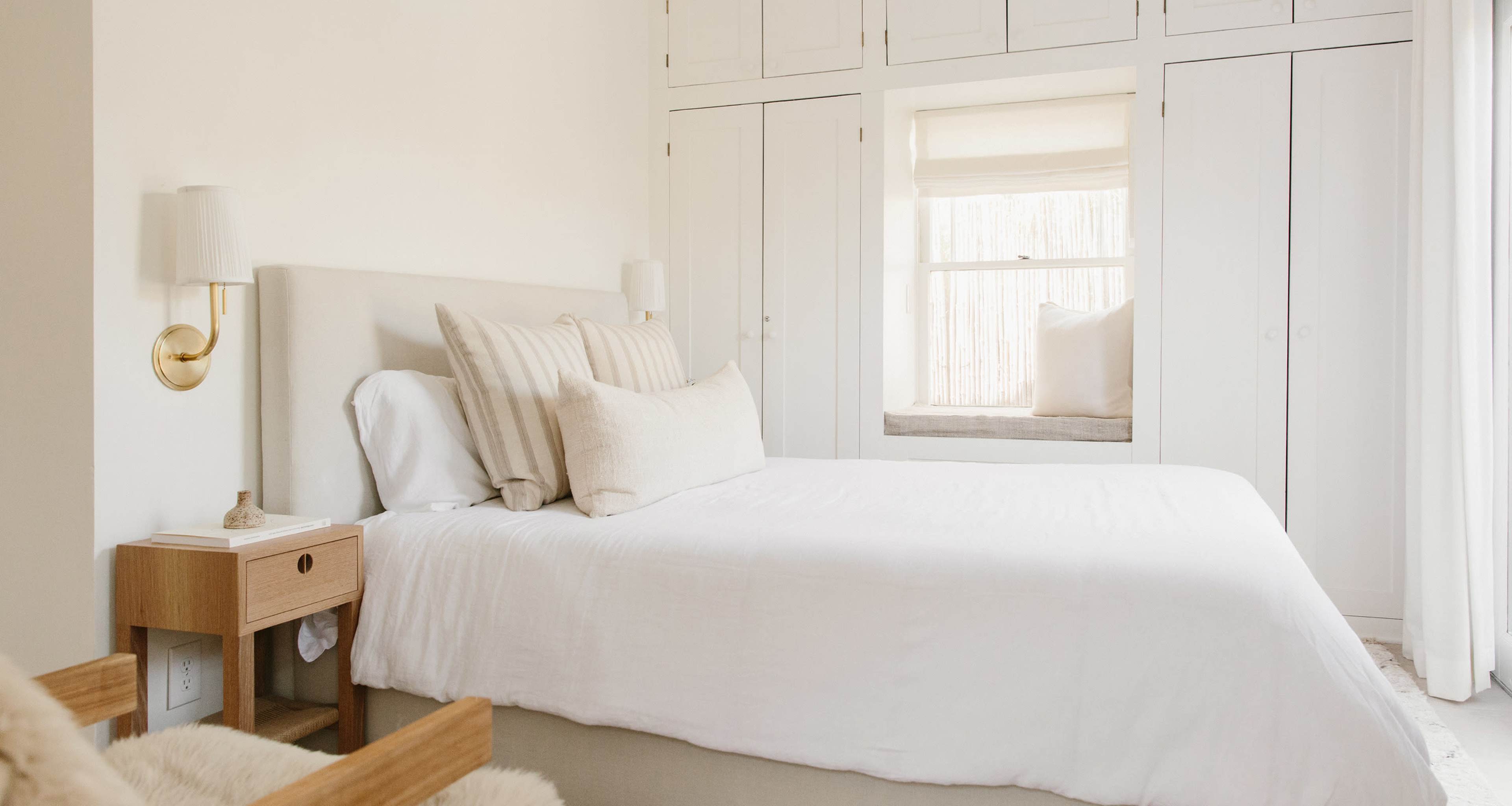A simple, well-lit bedroom with a white bedspread, several pillows, a wooden nightstand, and built-in cabinets, featuring a window seat.