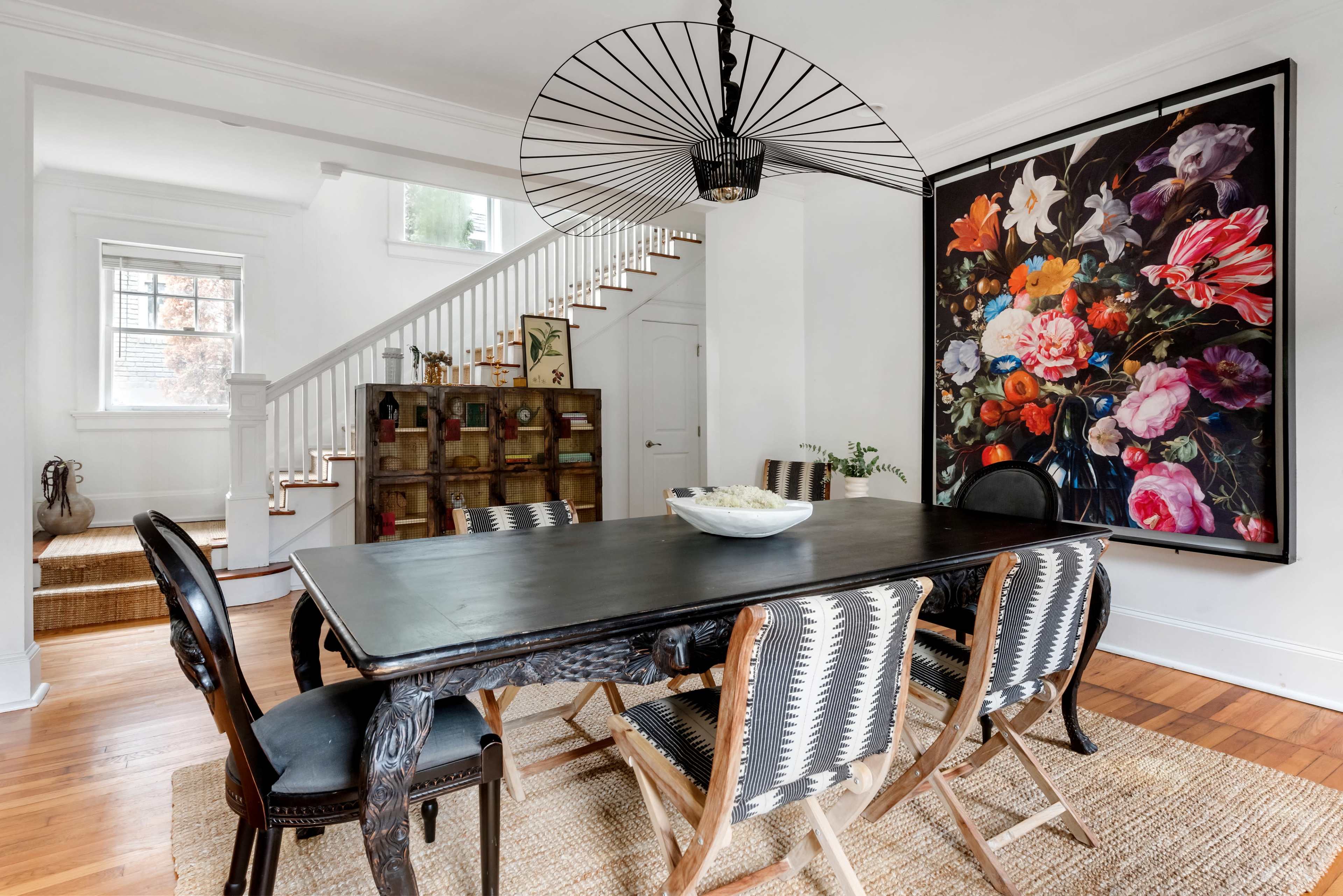 A dining room features a black table surrounded by six chairs, with a large floral artwork on the wall and a staircase in the background.