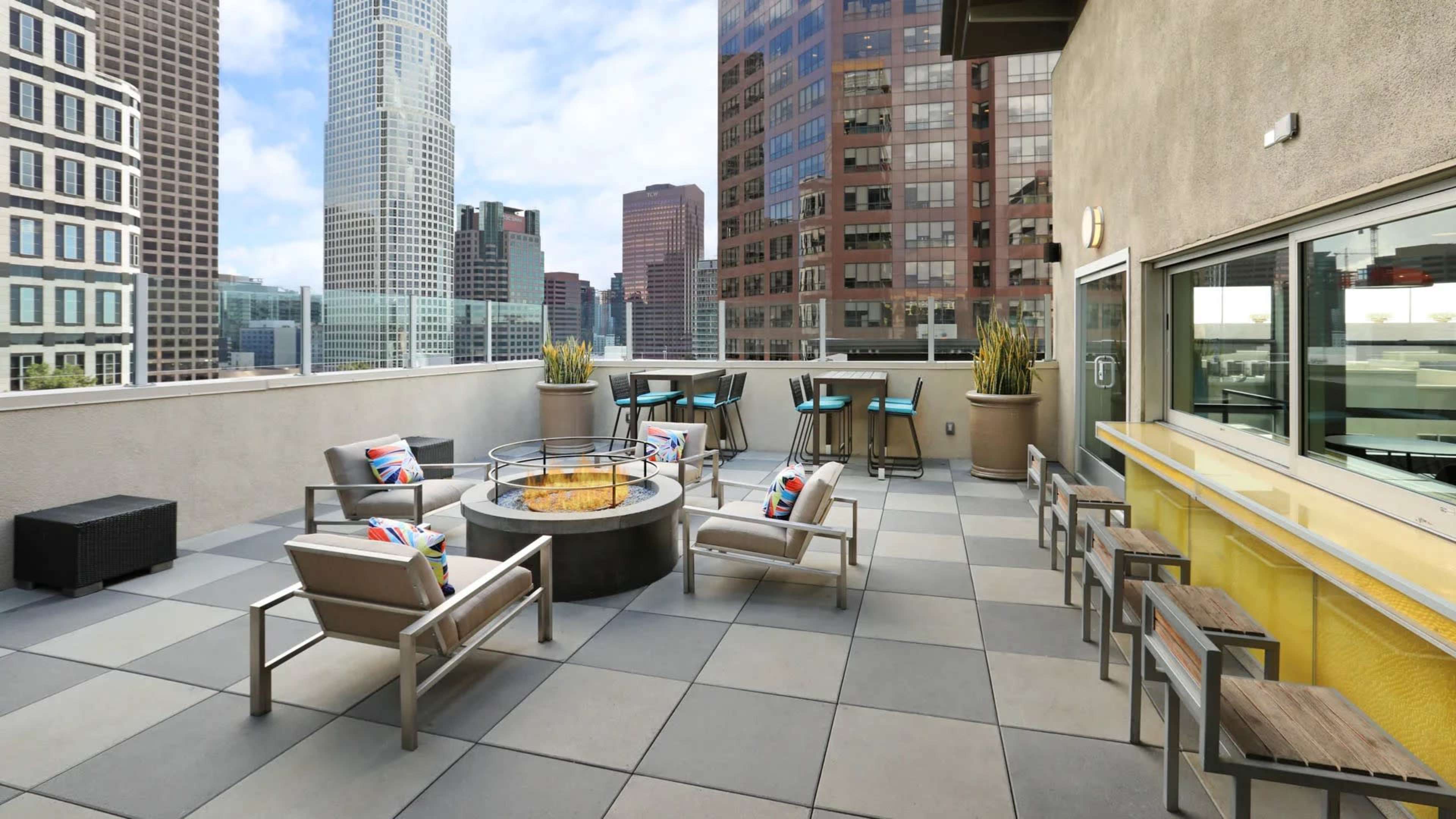 The image shows a rooftop terrace with seating arrangements around a fire pit, overlooking a city skyline.