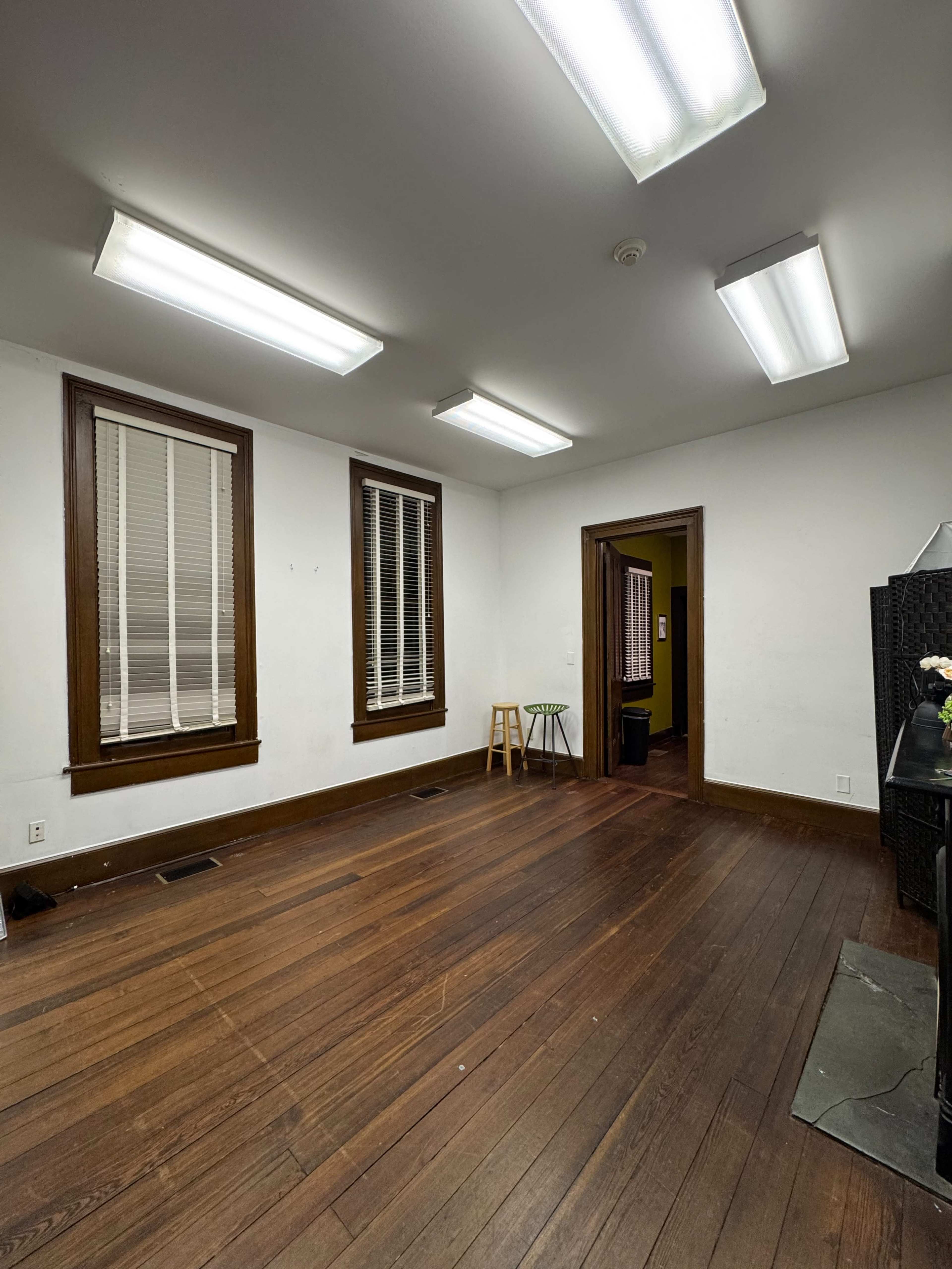 The image shows a sparsely furnished room with wooden flooring, illuminated by fluorescent ceiling lights, featuring two windows with blinds and an open doorway leading to another area.