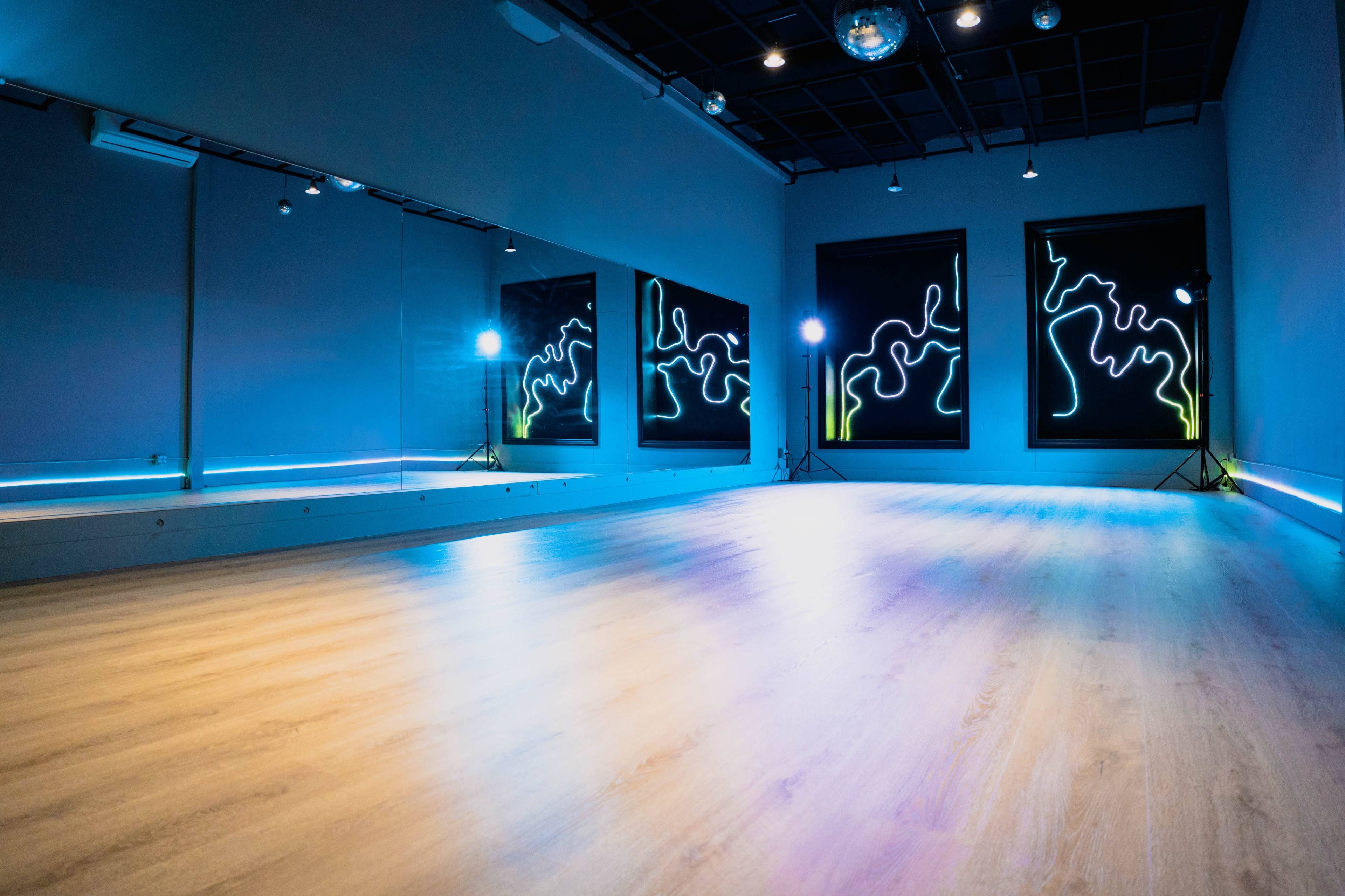 The image shows a dance studio with a smooth wooden floor, mirrors on one wall, and neon light artworks illuminating the space.