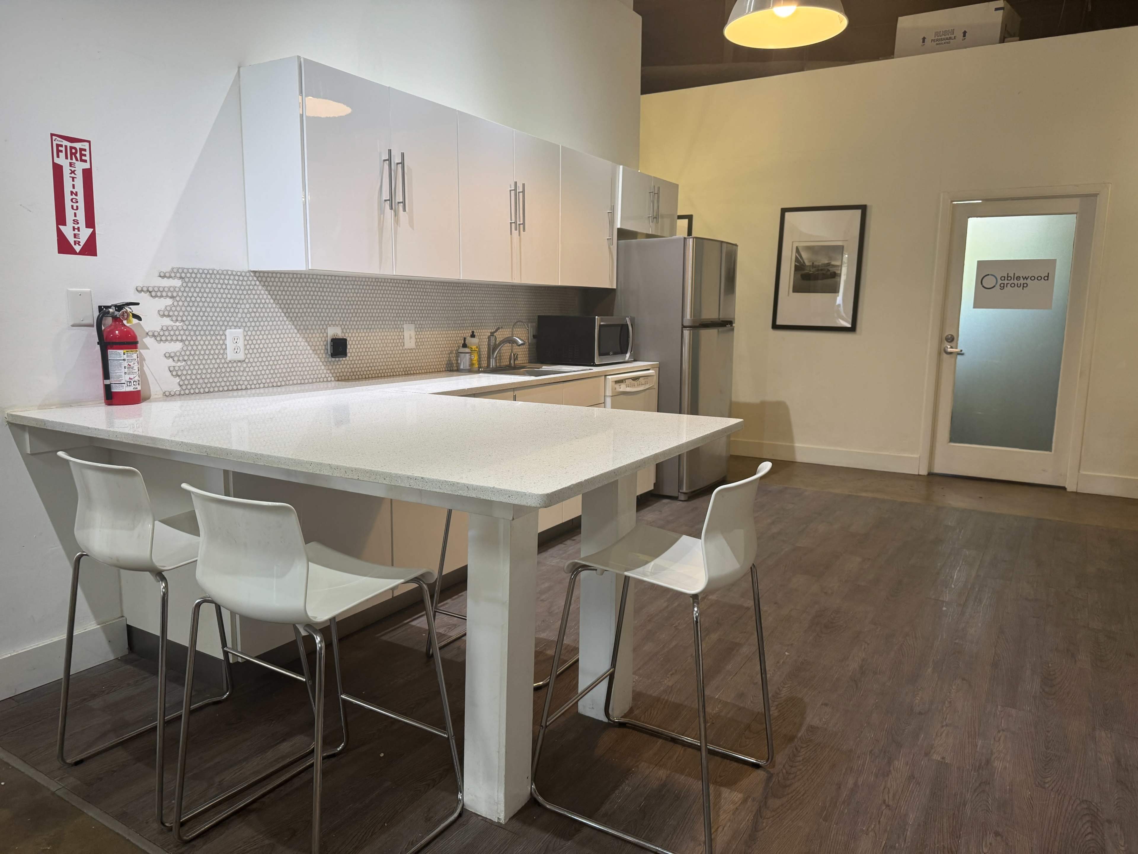 The image shows a modern kitchen with white cabinets, a white countertop, stainless steel appliances, and a dining area featuring three white bar stools.