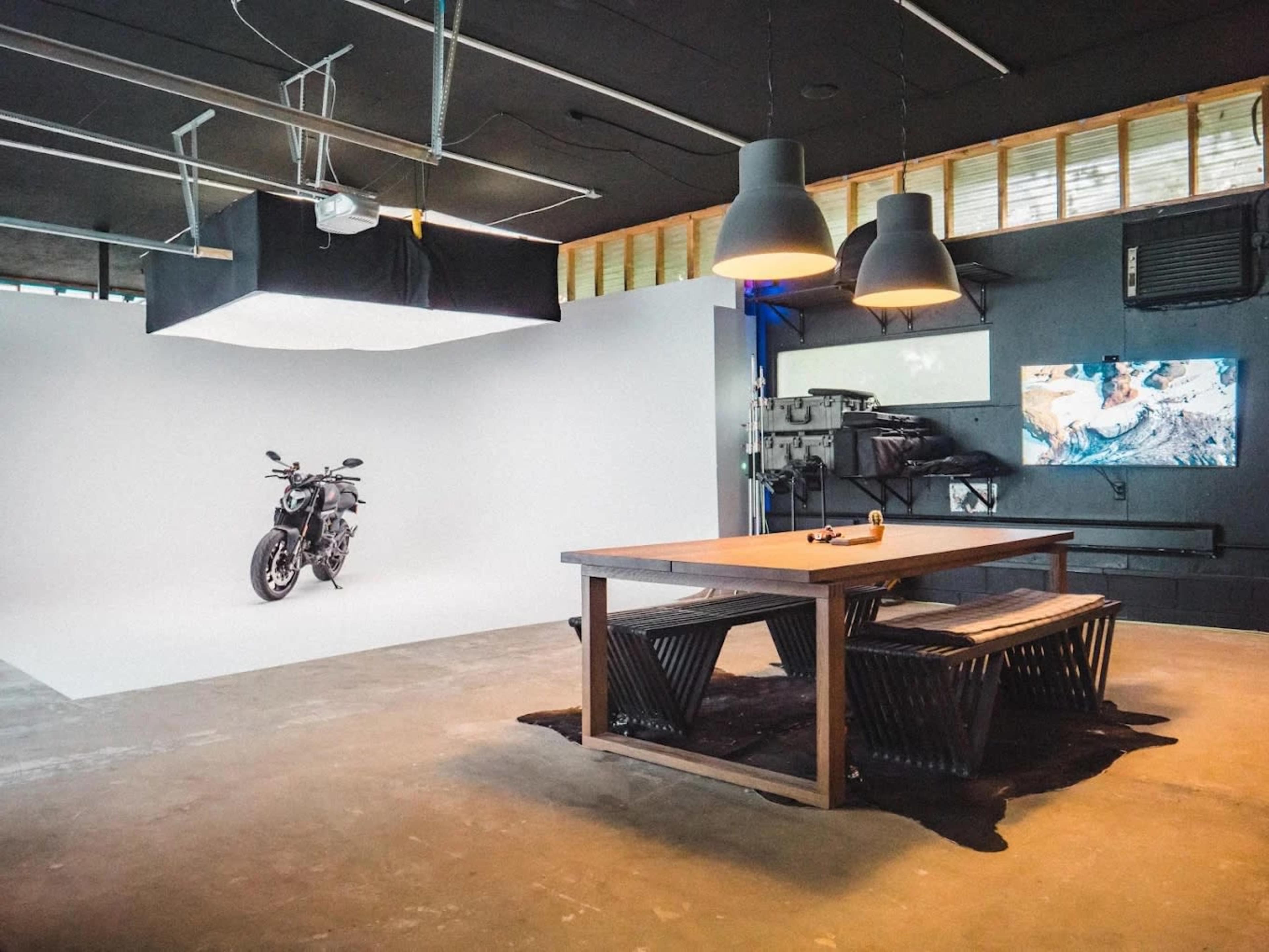 The image shows a minimalist studio space featuring a motorcycle placed on a white backdrop, accompanied by a wooden table and large pendant lights overhead.