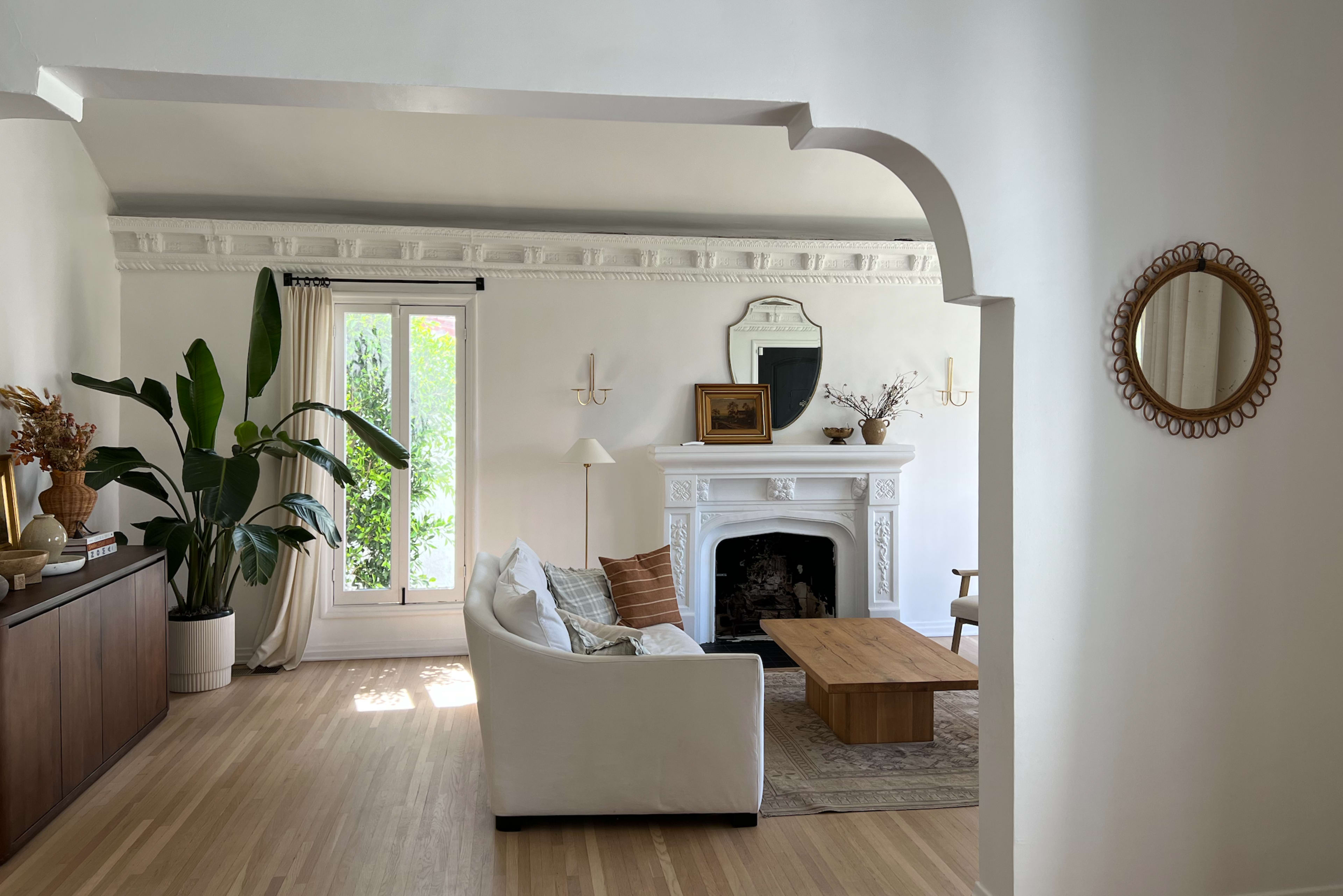 A living room features a white couch, a wooden coffee table, a fireplace with a decorative mirror above it, and large windows allowing natural light to enter.