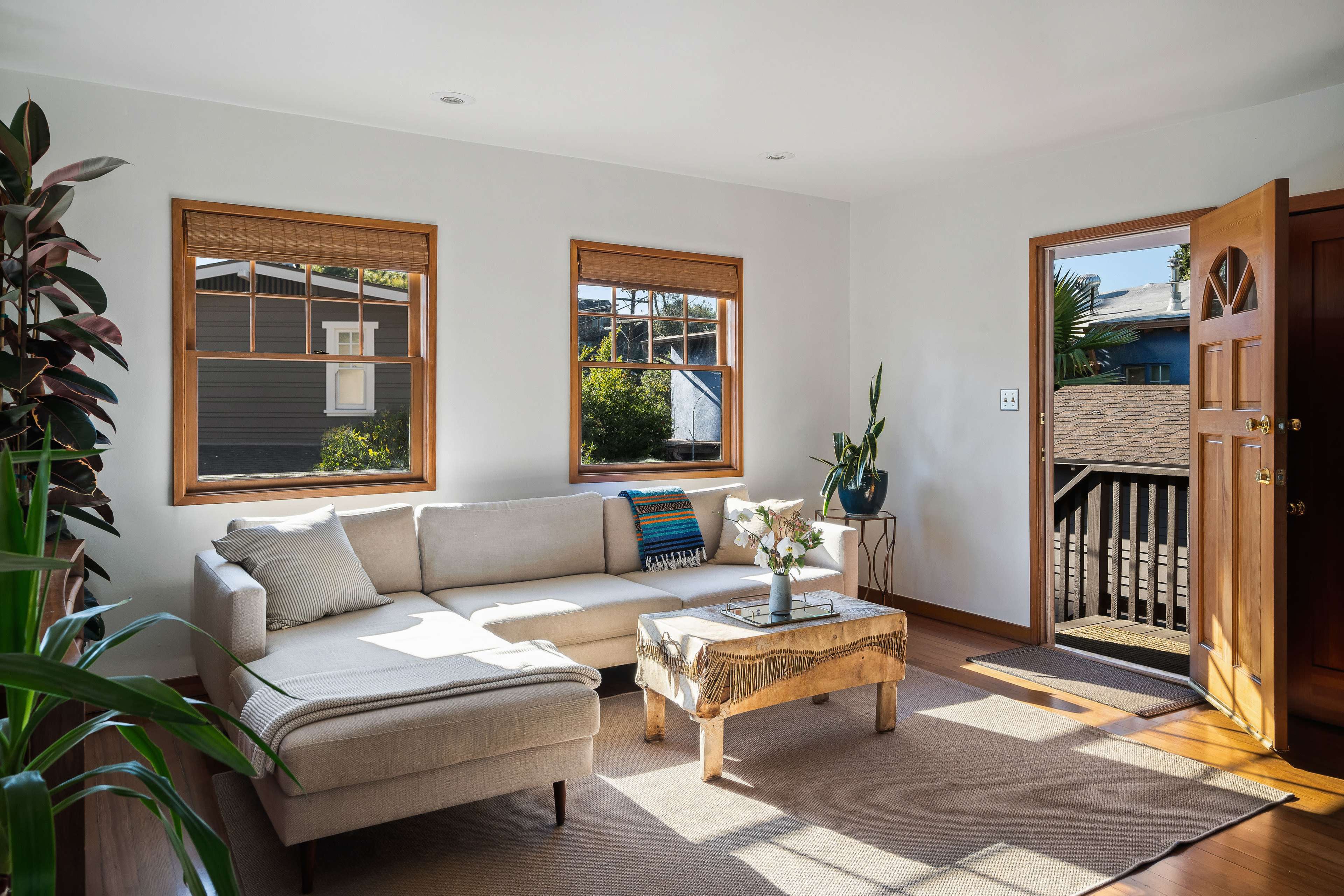 A light-filled living room features a sectional sofa, a coffee table, and three windows with views of the outside.