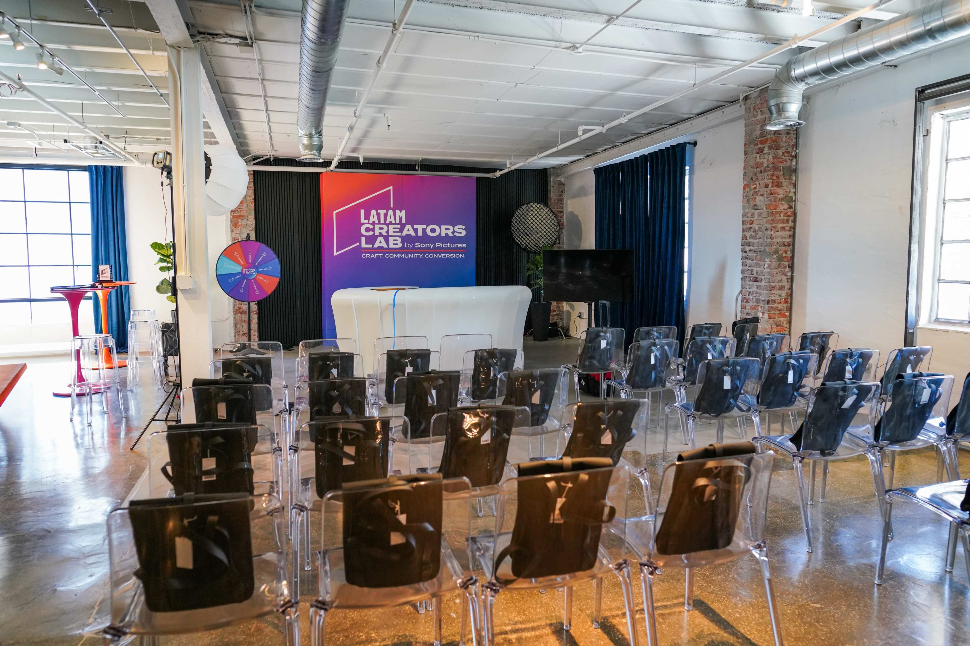 A spacious event room features rows of clear plastic chairs facing a stage with a colorful backdrop and various interactive elements like a spinning wheel.