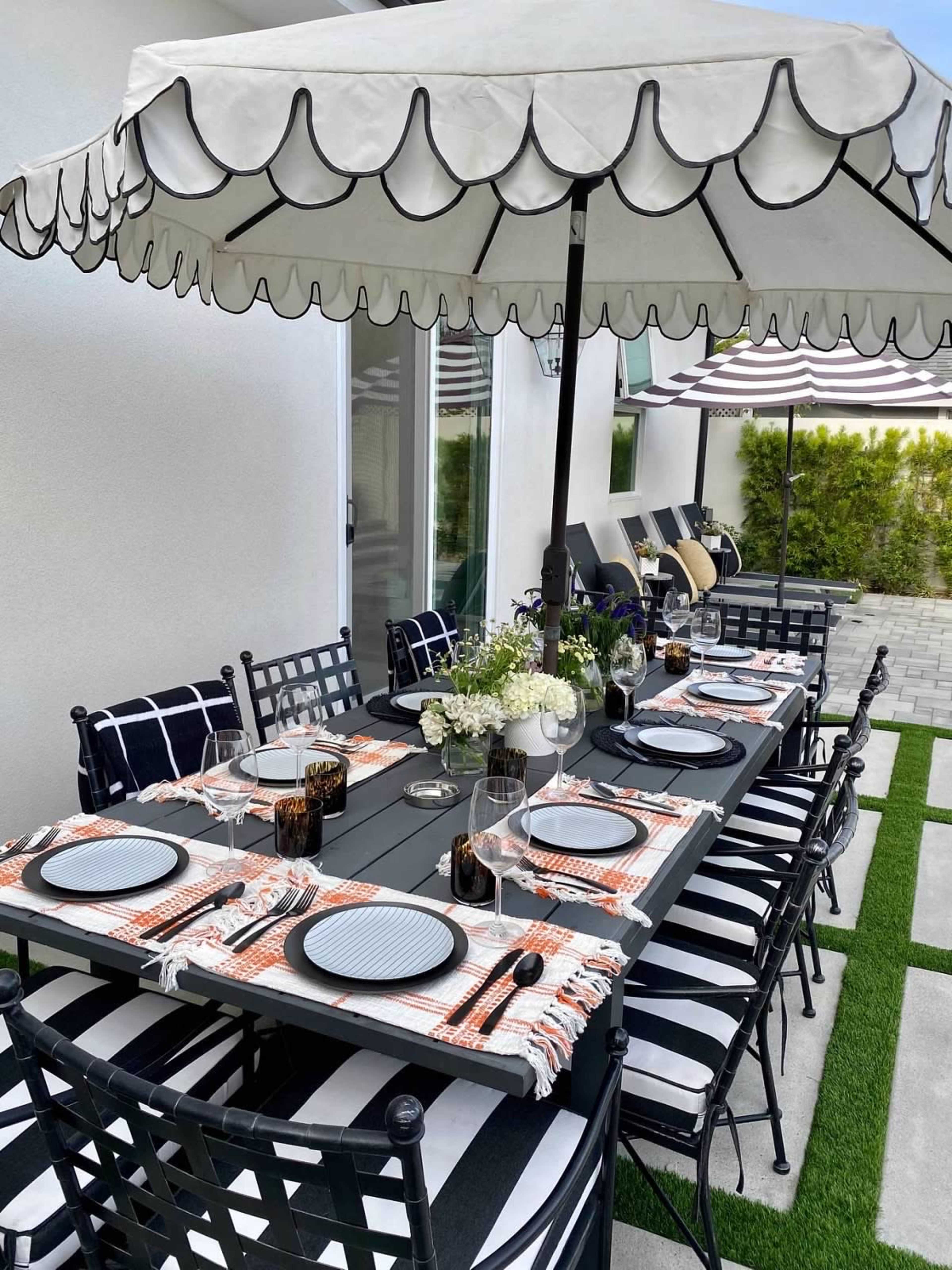 A large outdoor dining table is set with plates and glasses under a canopy umbrella, surrounded by black chairs on a patio with green grass.