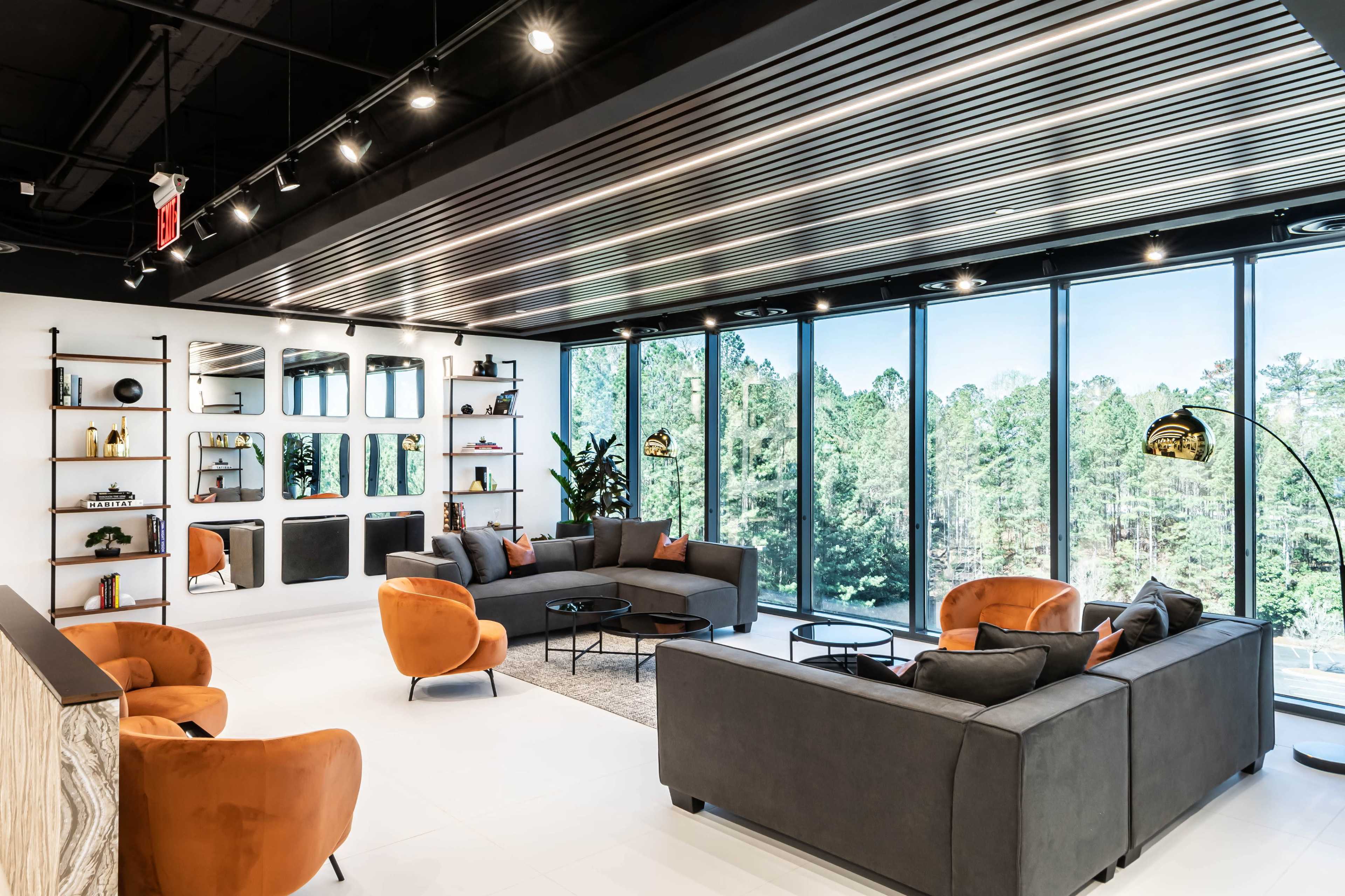 A modern lounge area features gray sofas and orange armchairs with large windows overlooking a green landscape.