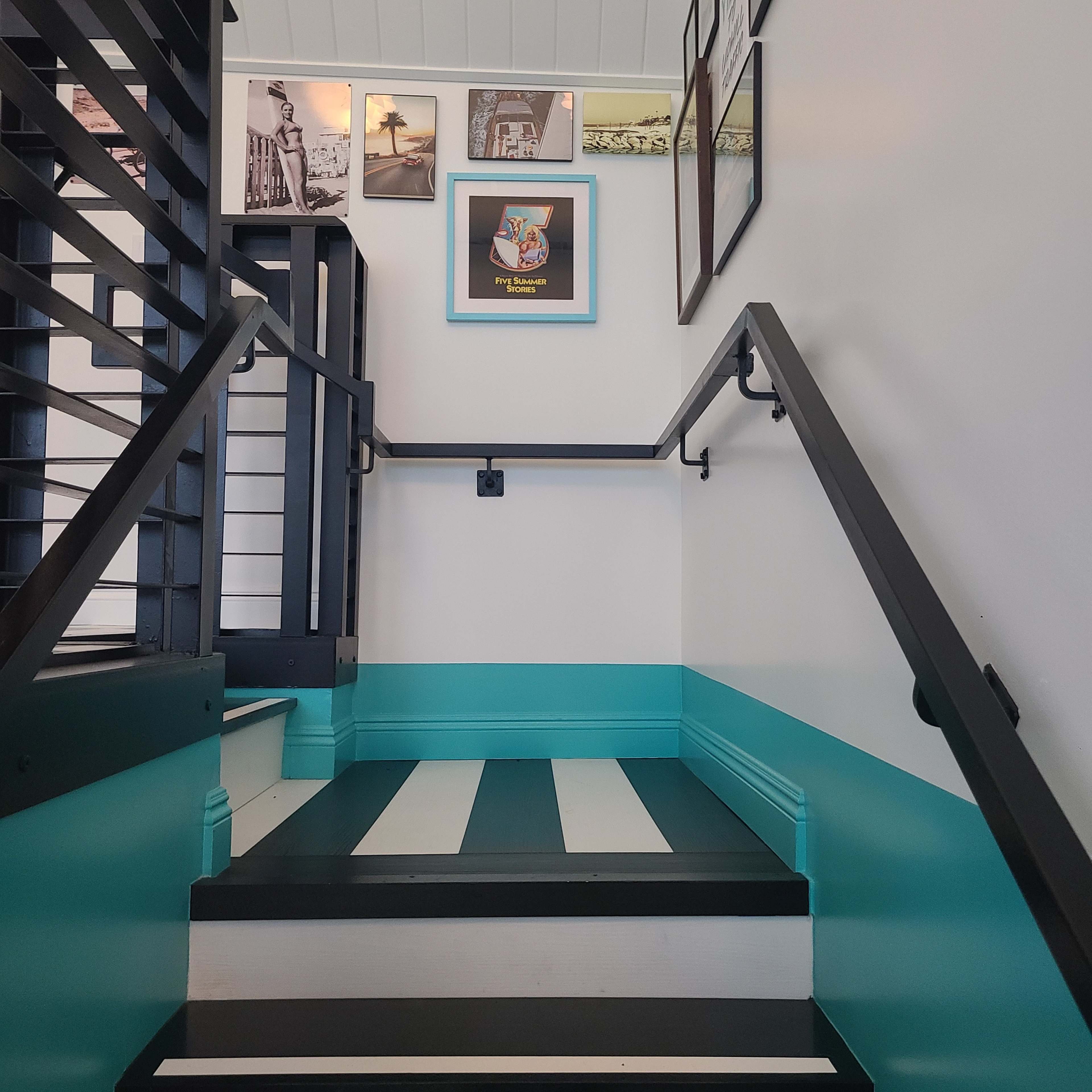 The image shows a stairway with striped floors and a turquoise wall, leading up to a gallery of framed photos.