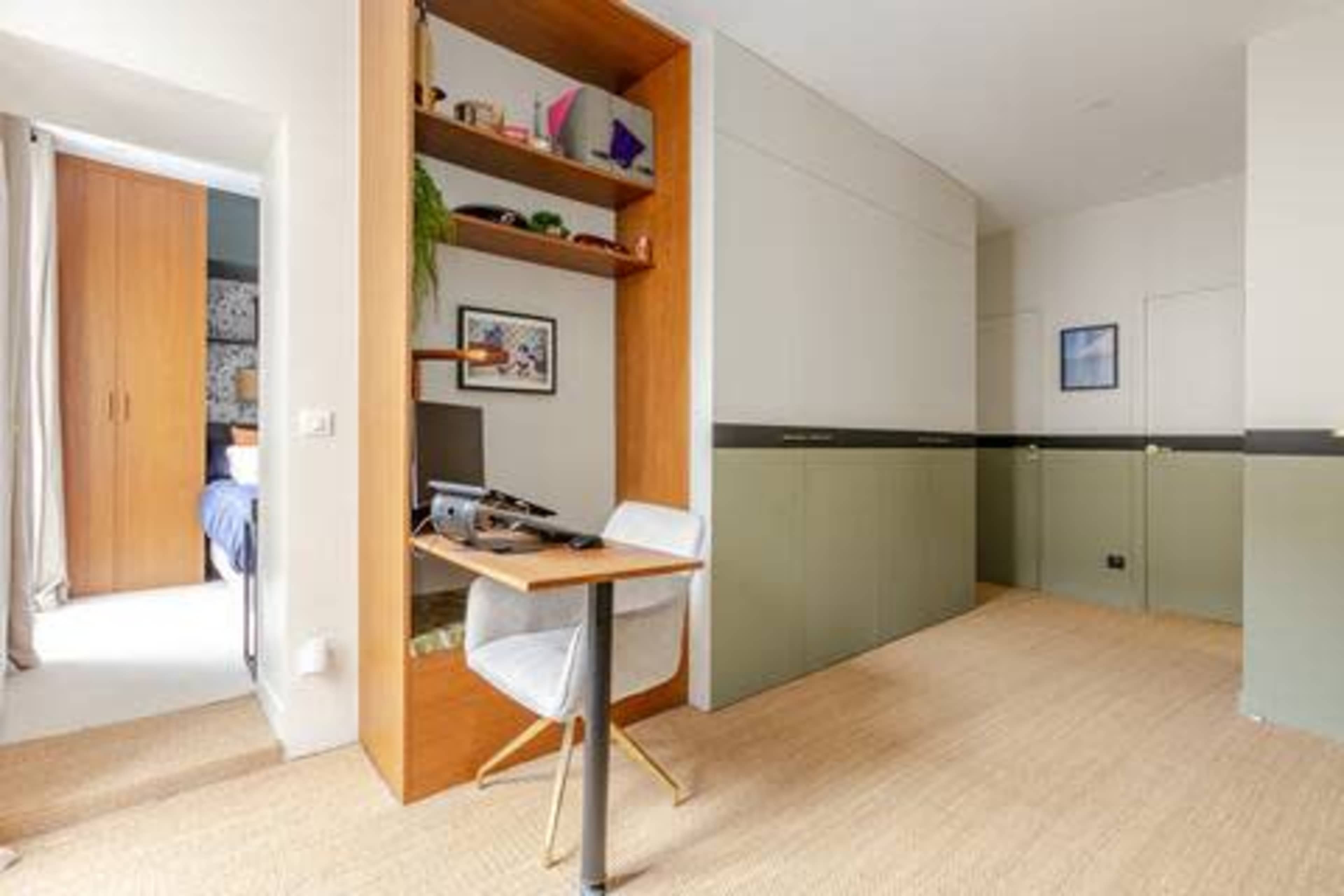 The image shows a modern interior space with a small desk and computer setup against a wall, and a separate area with a bed visible in the background.