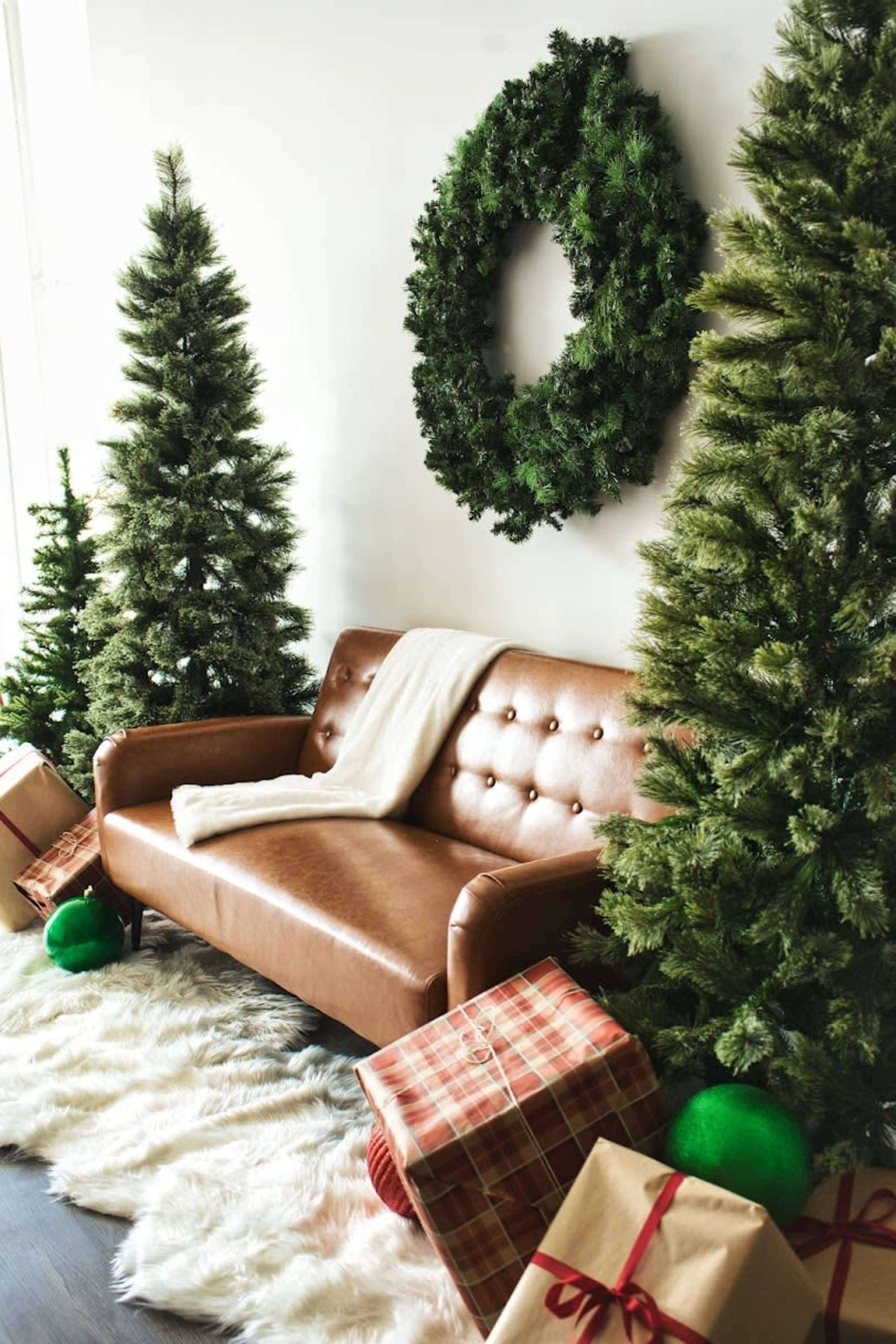 A brown leather sofa is flanked by two Christmas trees and decorated with a large wreath on the wall, surrounded by wrapped gifts.