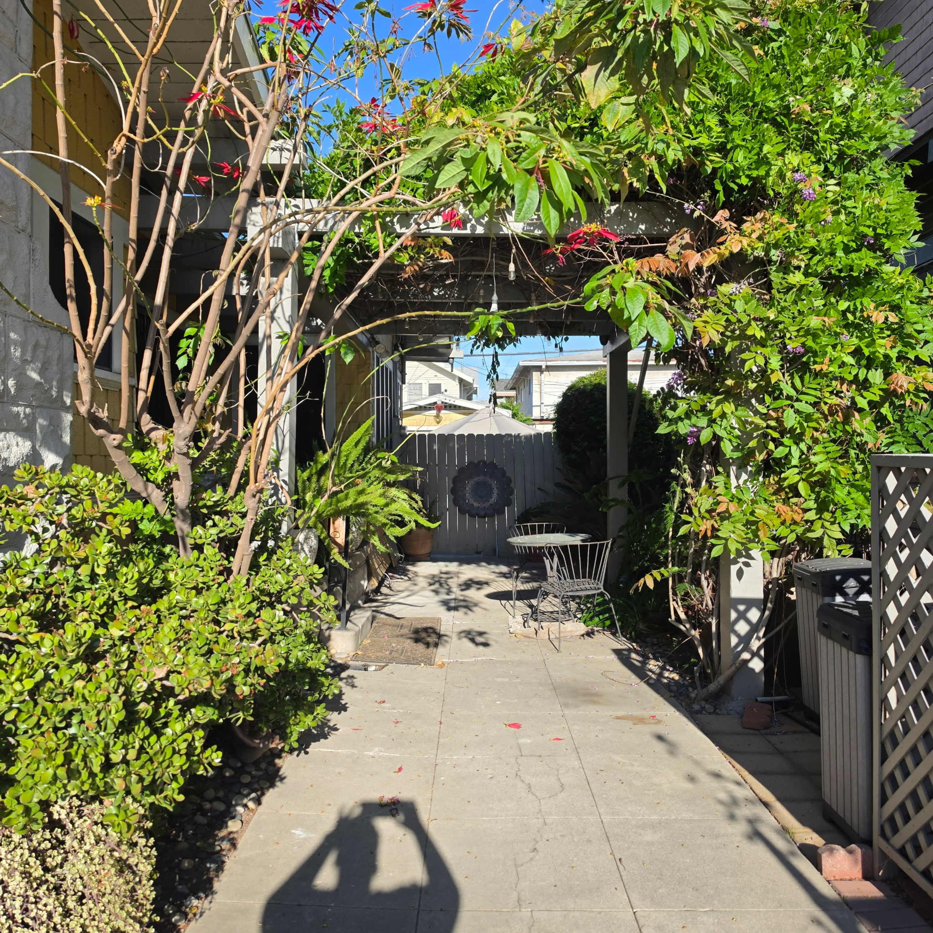 The image shows a narrow, landscaped walkway lined with plants and a wooden fence, leading to a small patio area with a table and chairs.