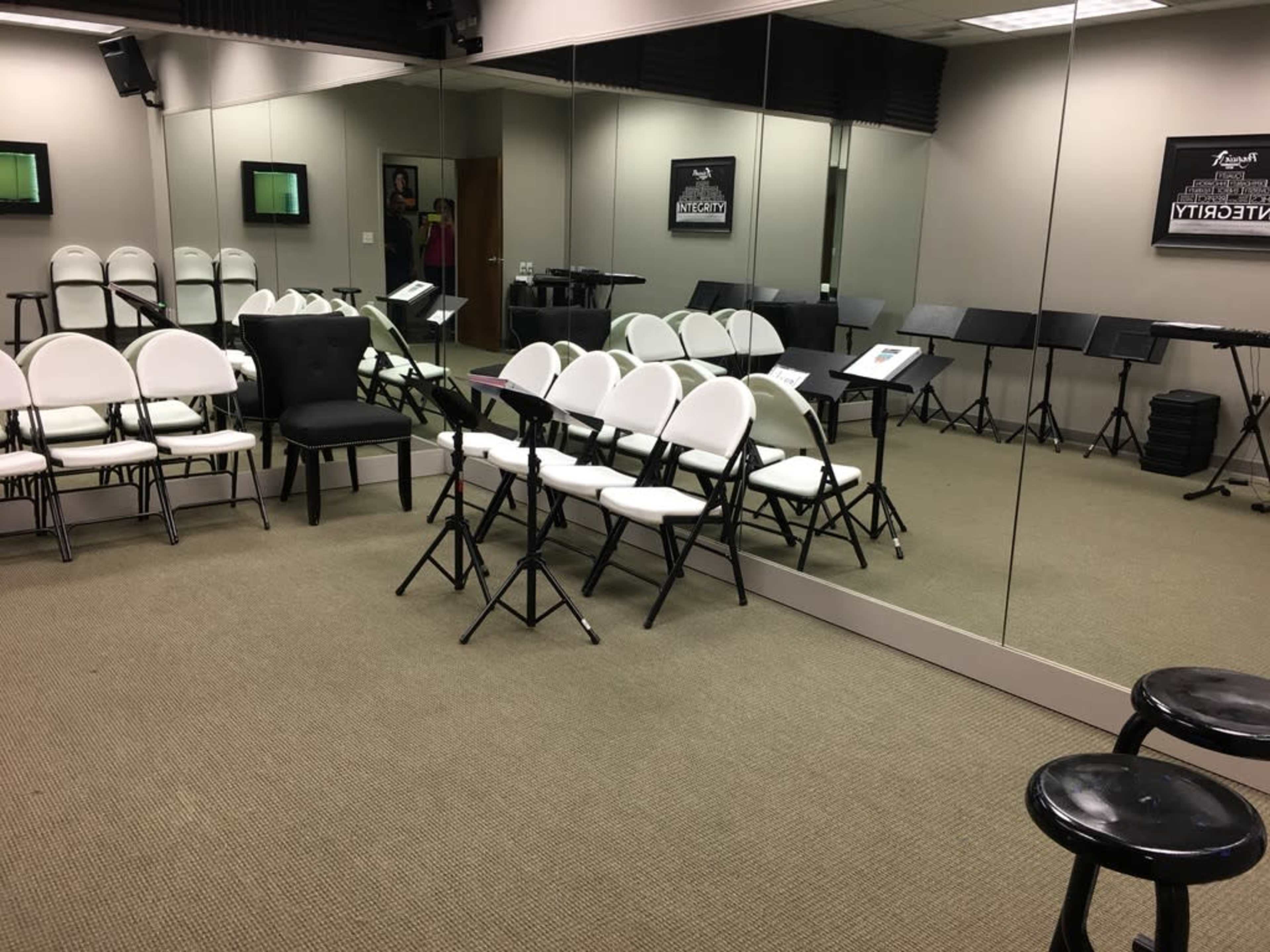 A music rehearsal room features several rows of white and black chairs, music stands, and large mirrors on the walls.