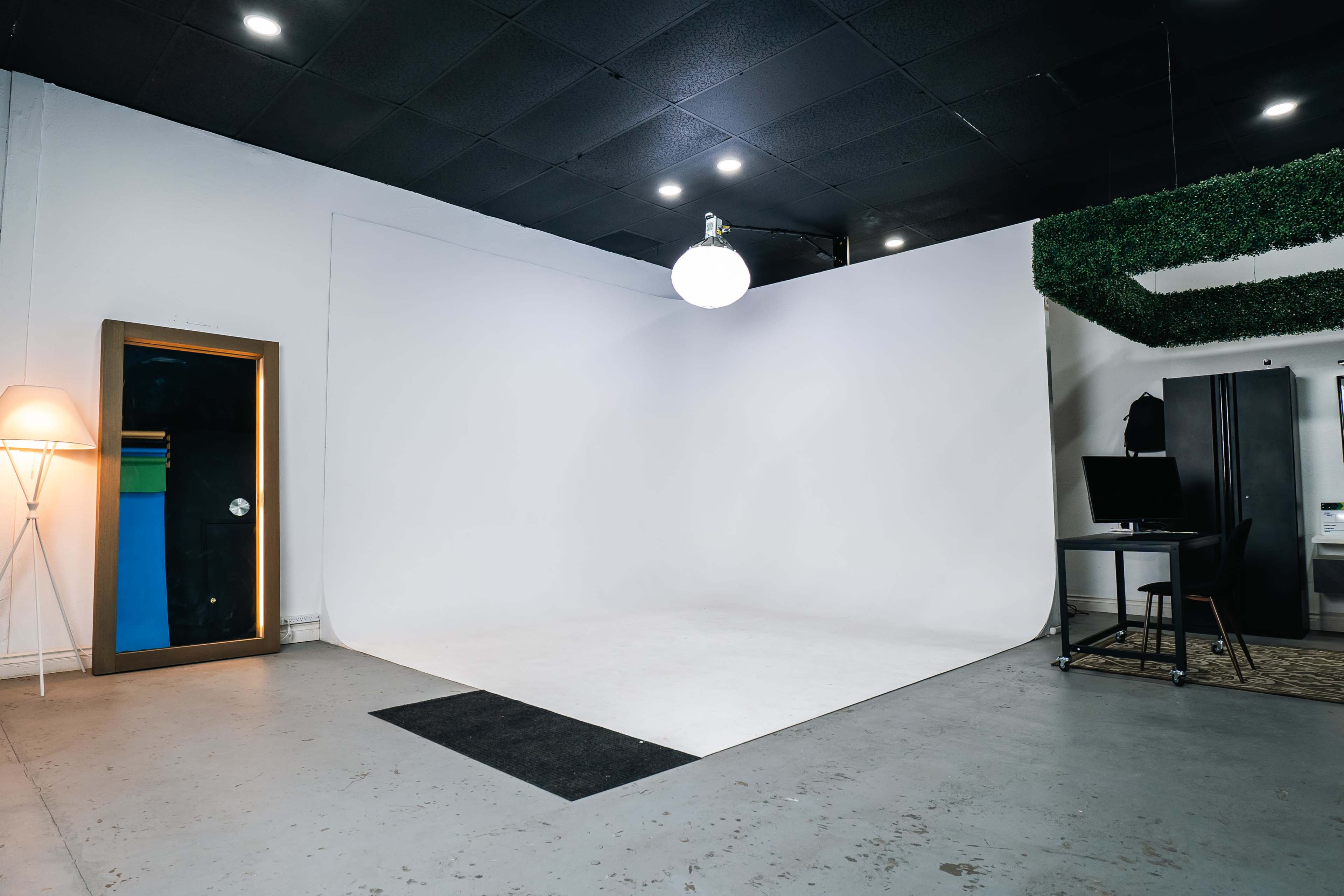 A minimalist room with a white backdrop, a circular ceiling light, a mirrored cabinet, a desk with a computer, and a green wall feature.