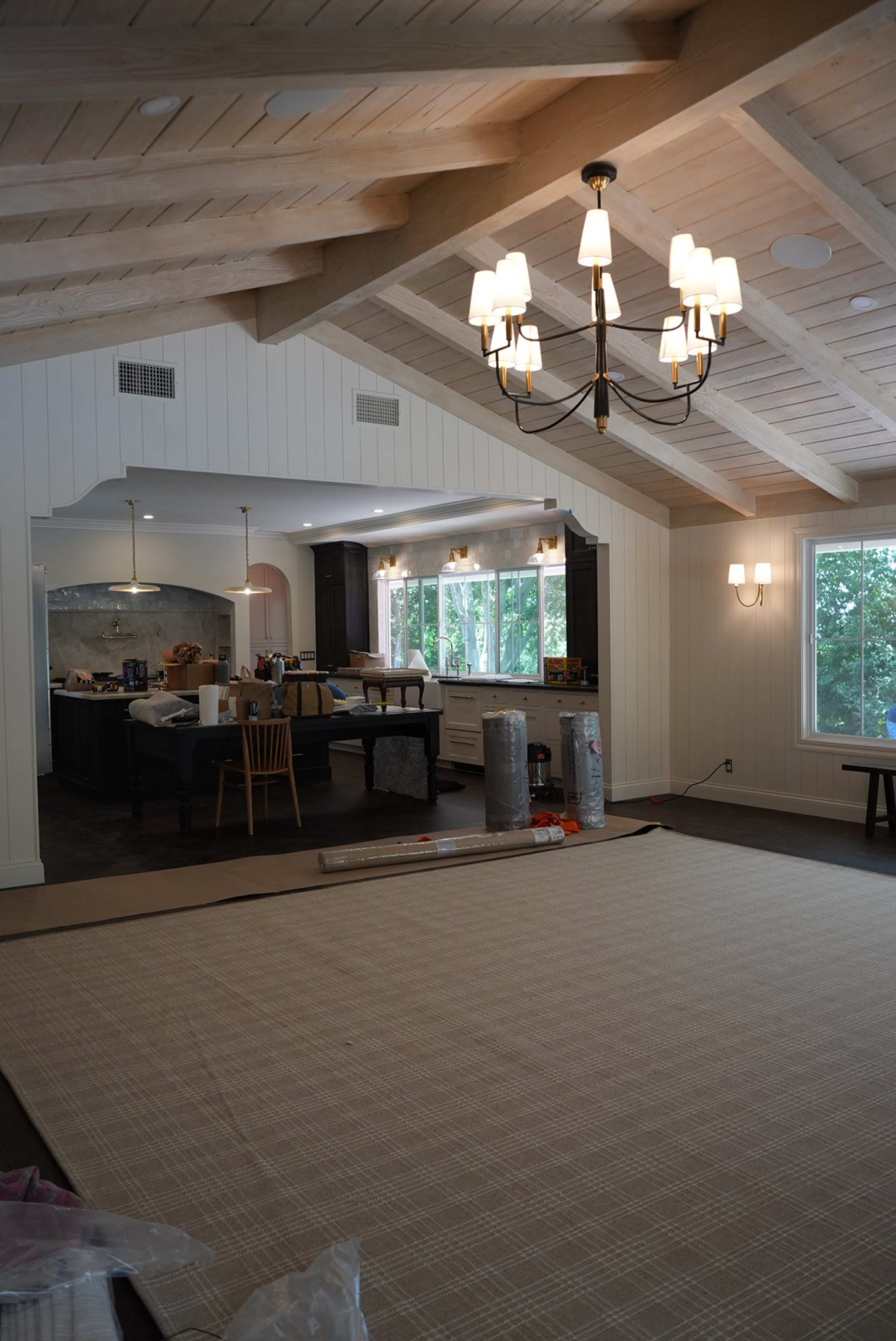 The image shows a spacious living area with a dining table, modern kitchen, and large windows, featuring exposed wooden beams and a patterned rug on the floor.