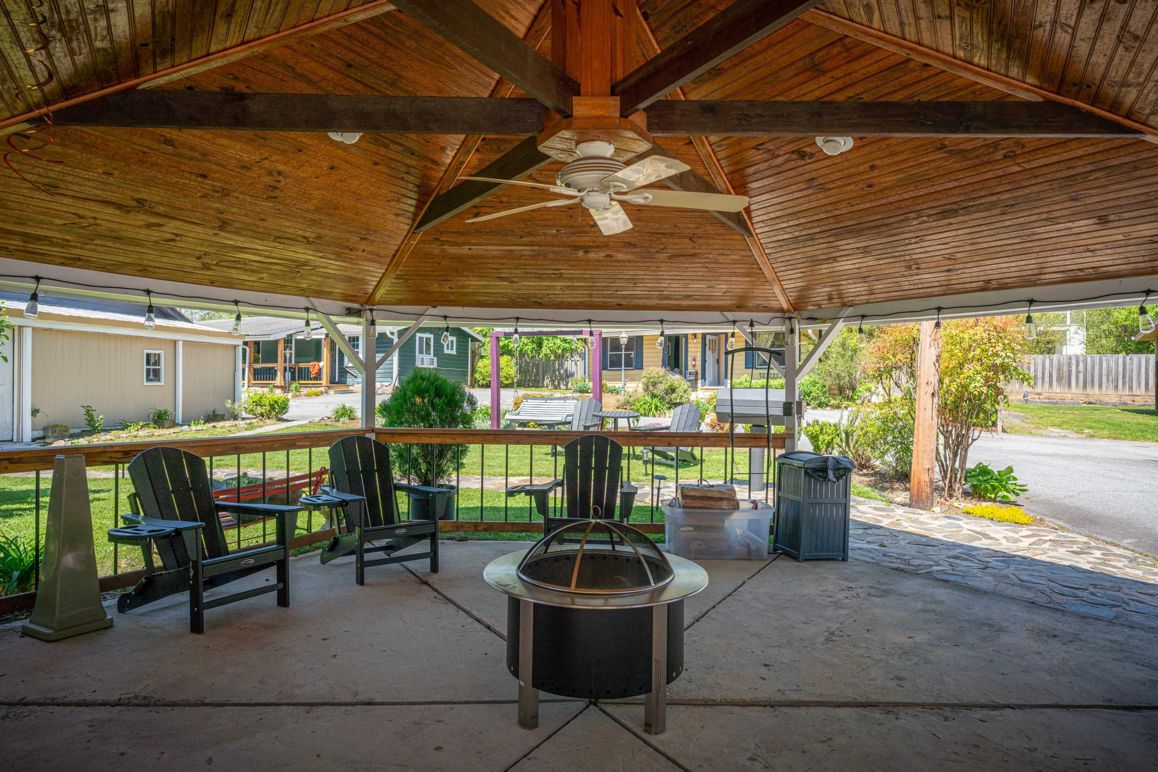 A covered gazebo with wooden beams, a ceiling fan, and four black Adirondack chairs arranged around a central fire pit.