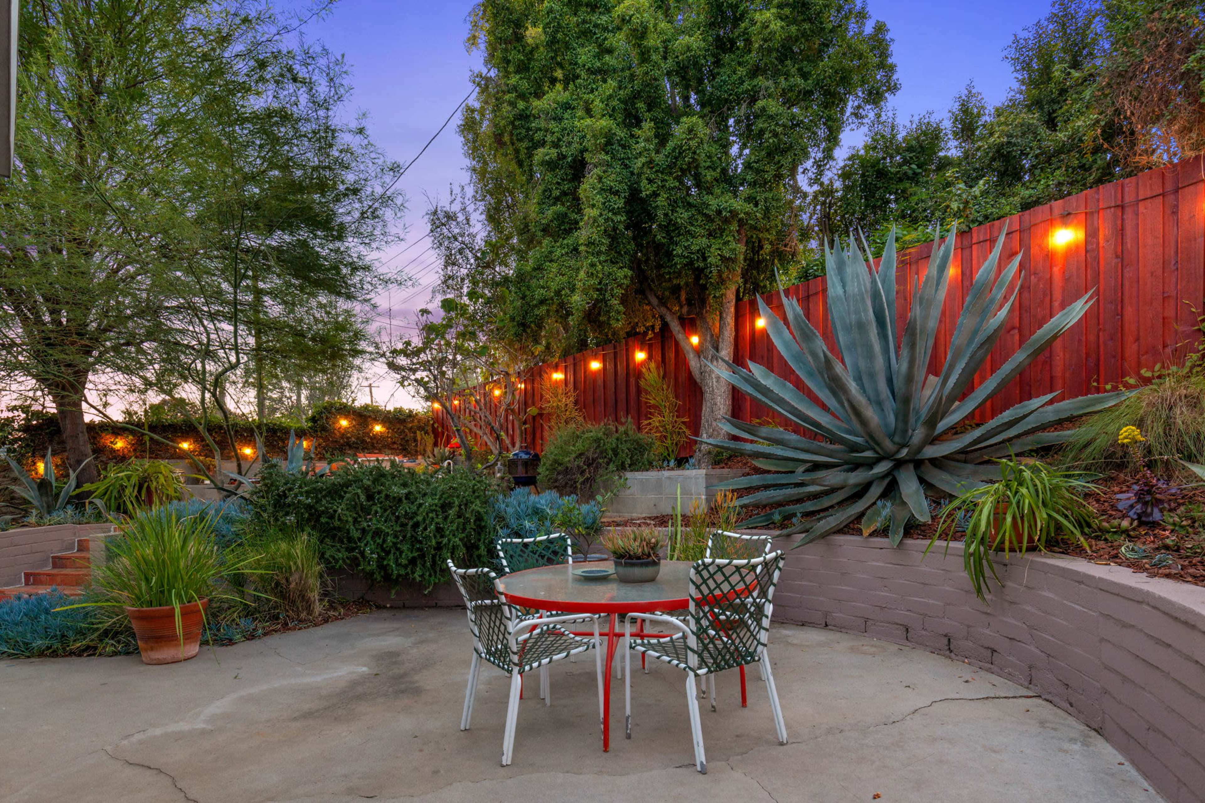 A small outdoor seating area features a round table surrounded by chairs, with a large agave plant and decorative lighting along a wooden fence in the background.