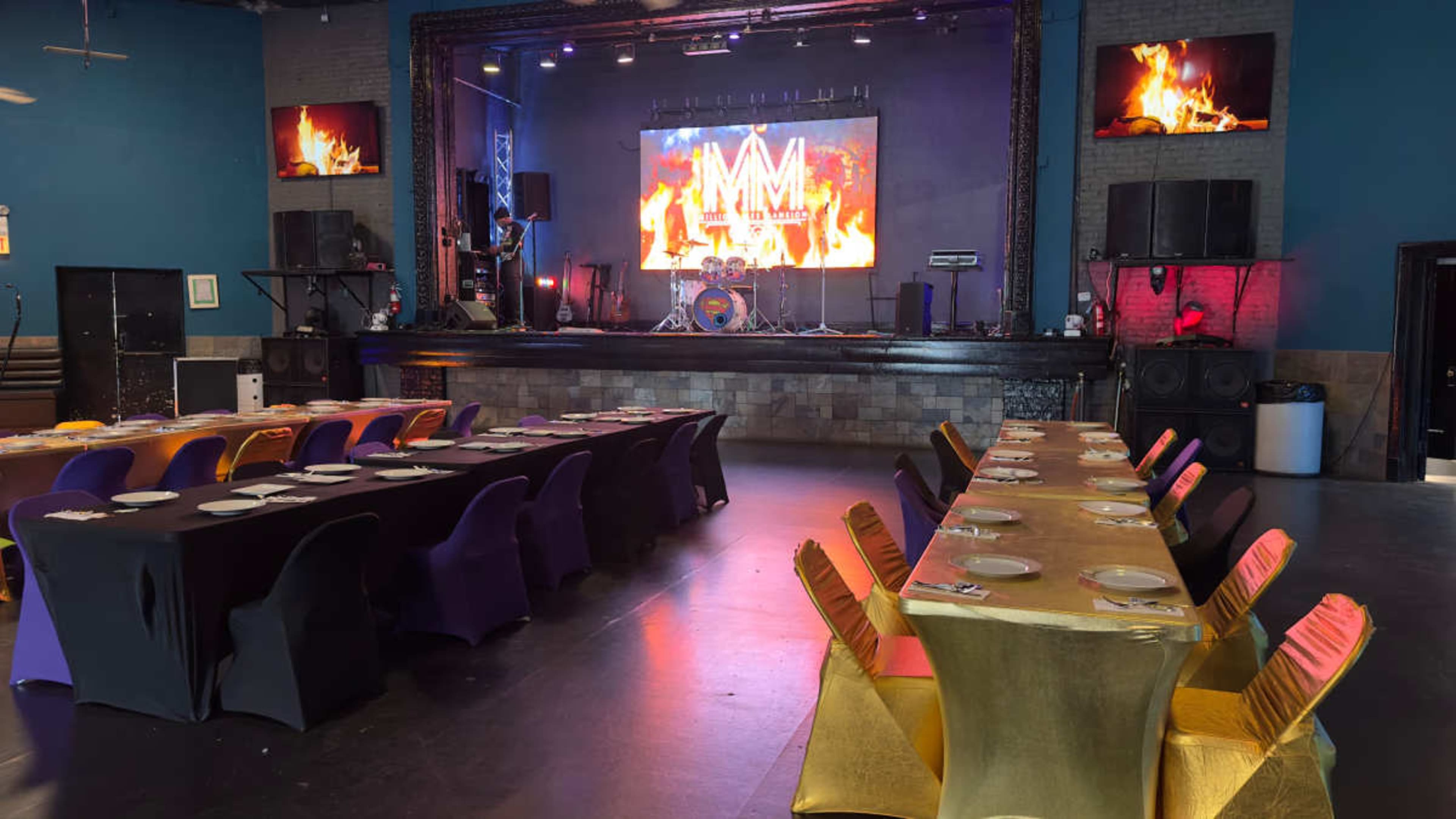 A stage with a large display is set behind rows of tables covered in colorful tablecloths in a venue designed for performances and events.