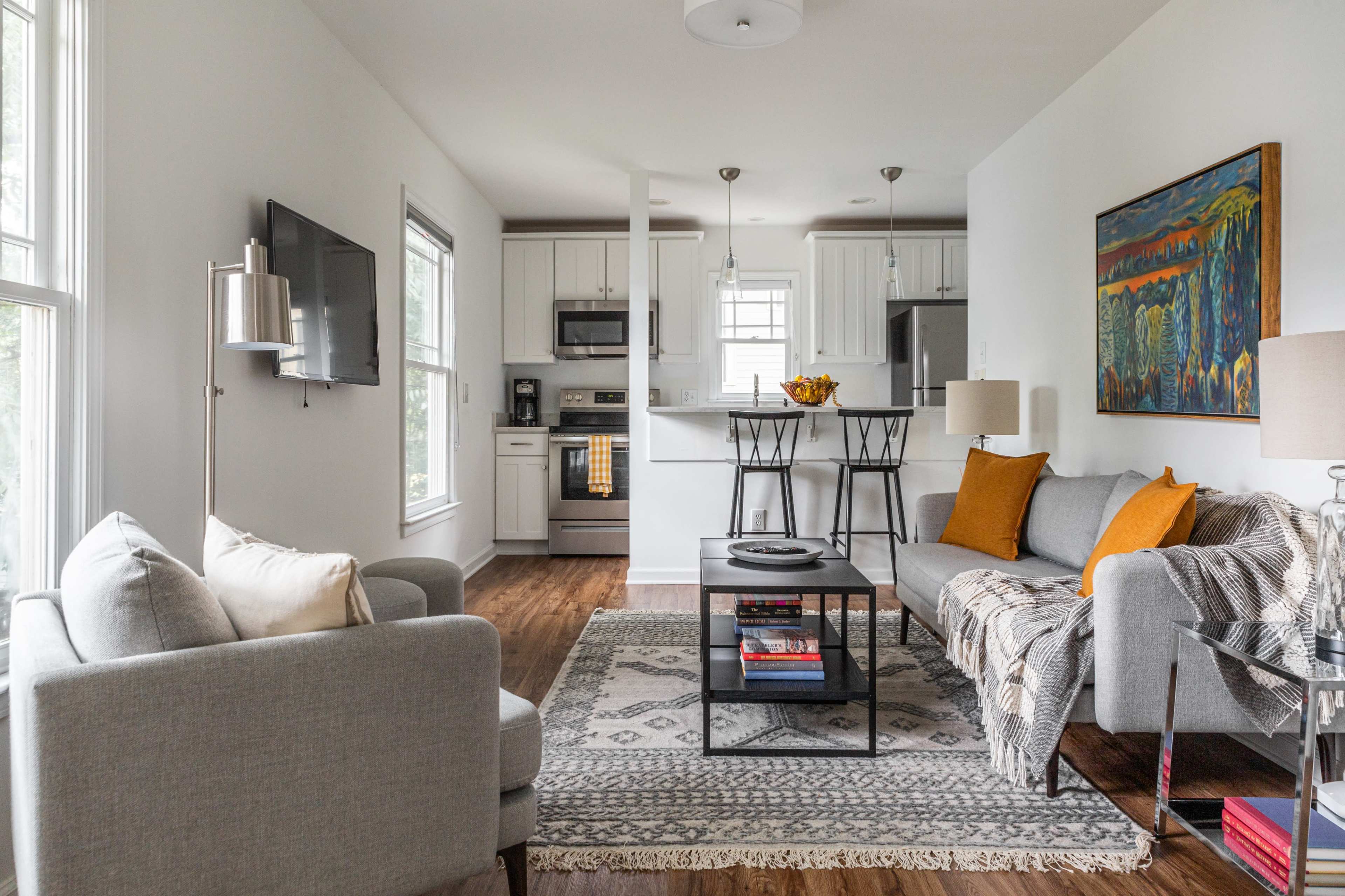 A bright living area featuring a gray sofa, an accent chair, a coffee table with books, and a kitchenette in the background.