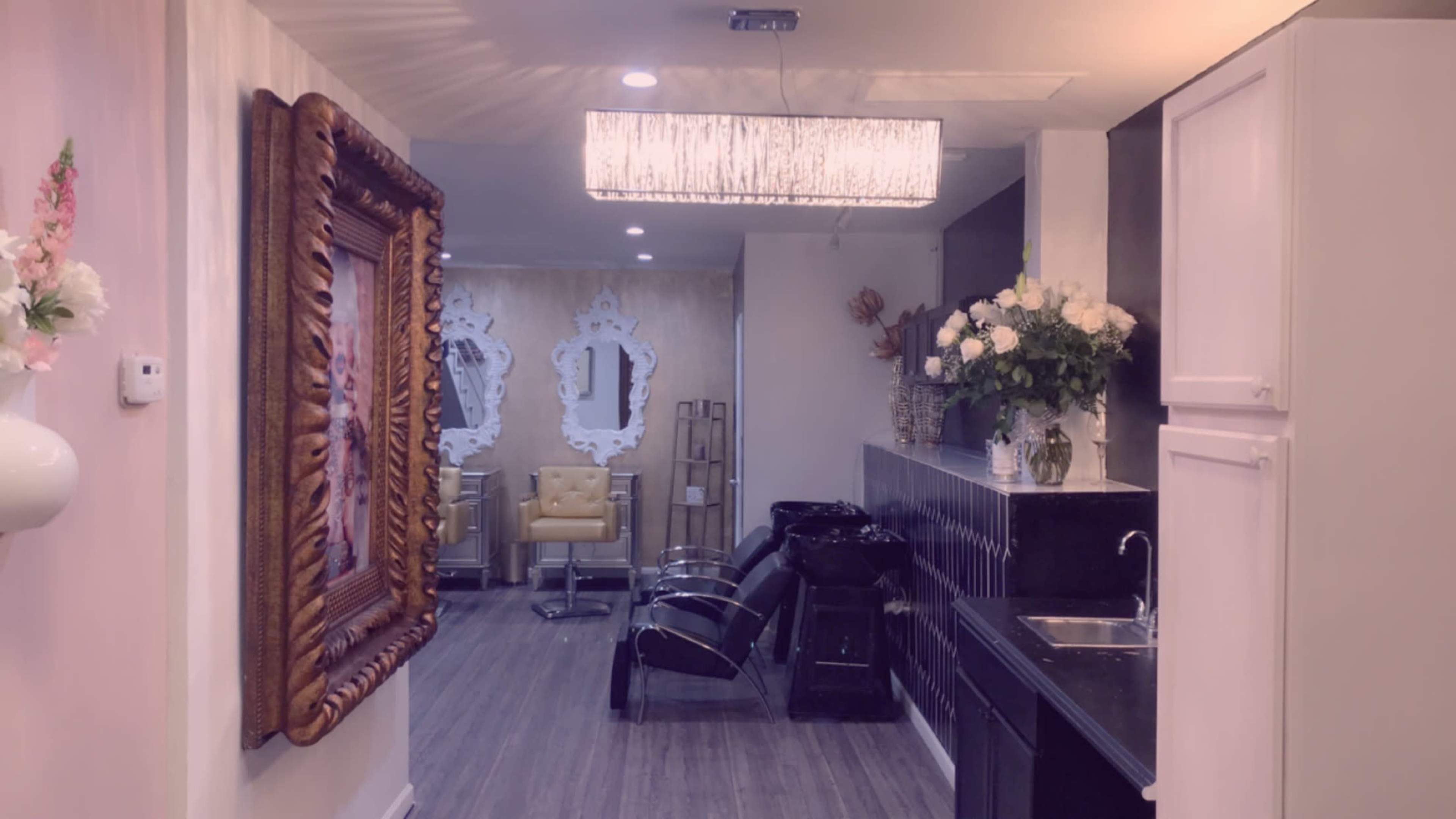 A corridor in a salon featuring a large decorative mirror, hair-washing stations, and stylish chairs, with floral arrangements and ambient lighting.