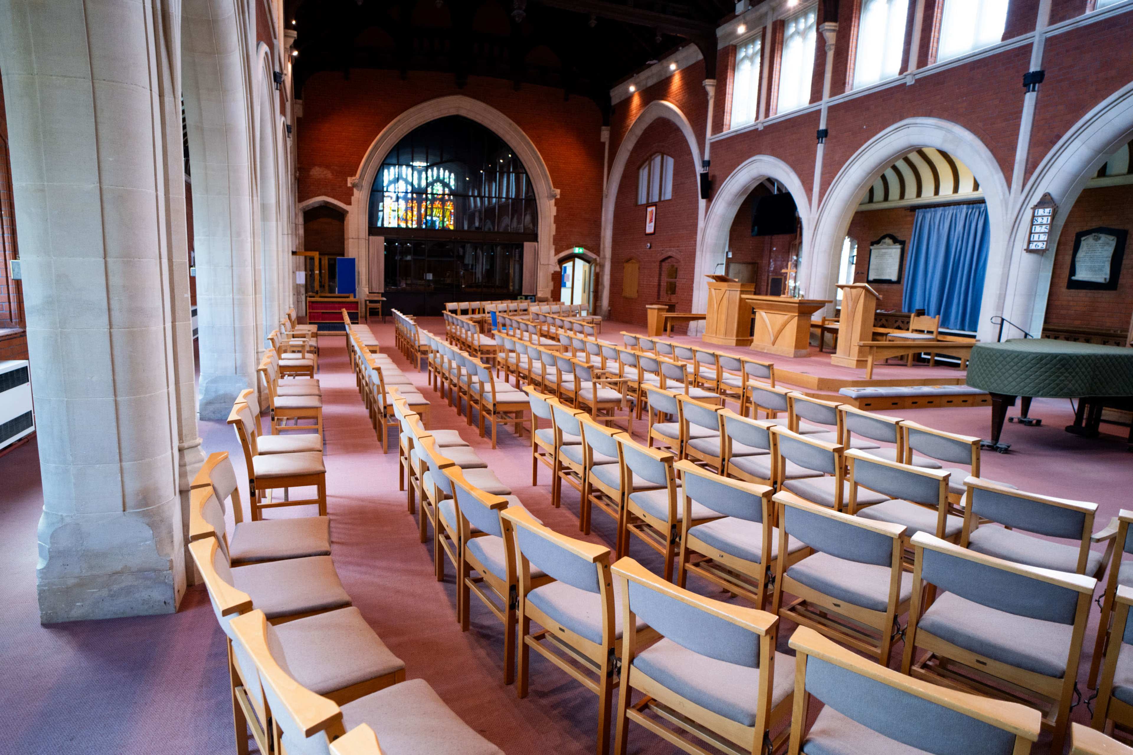 The image shows a spacious interior of a hall with rows of wooden chairs arranged facing a podium, featuring large arched windows and a stained glass design.