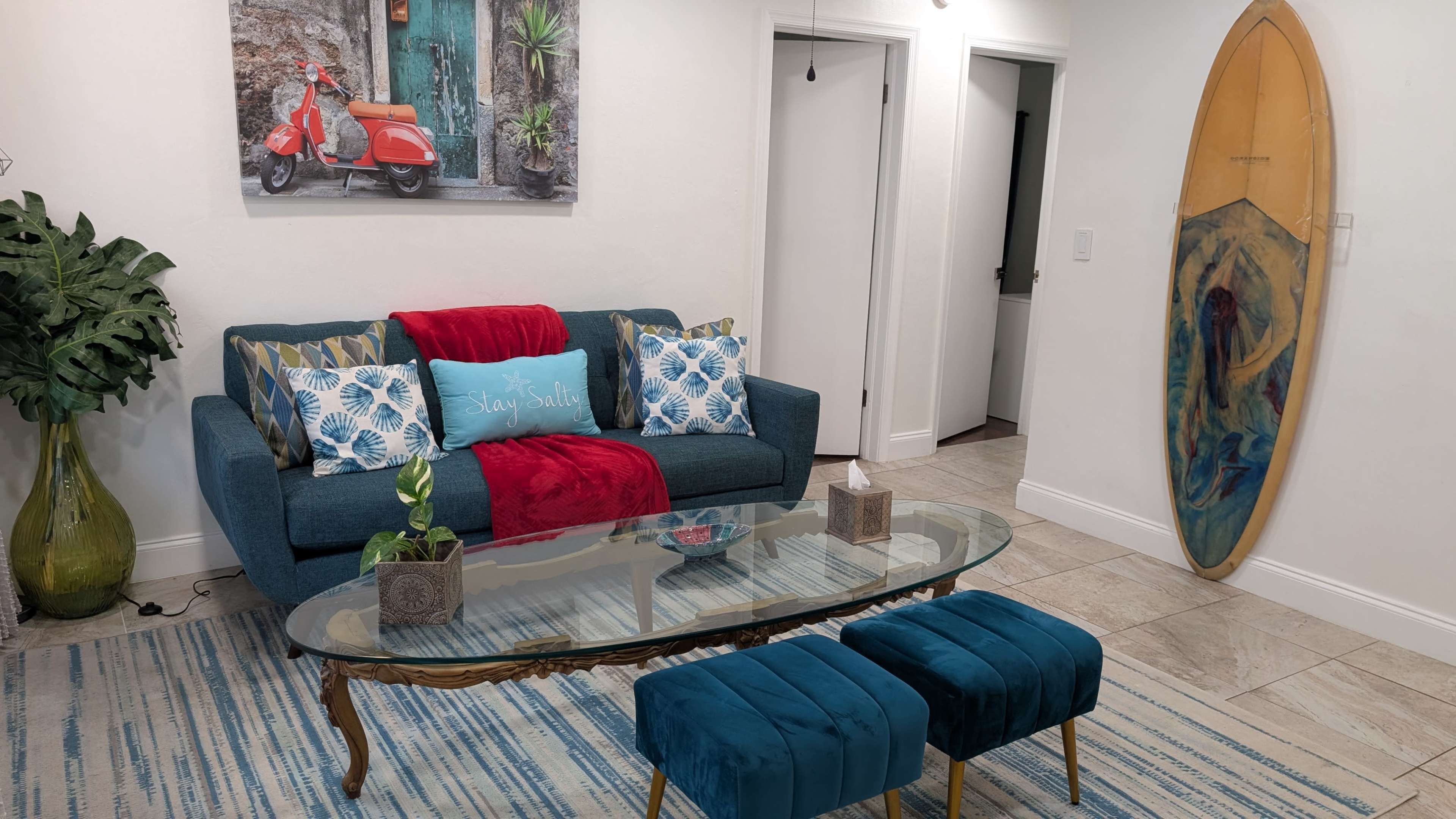 The image shows a living room featuring a blue sofa with decorative pillows, a glass coffee table, two blue ottomans, and a surfboard against the wall.