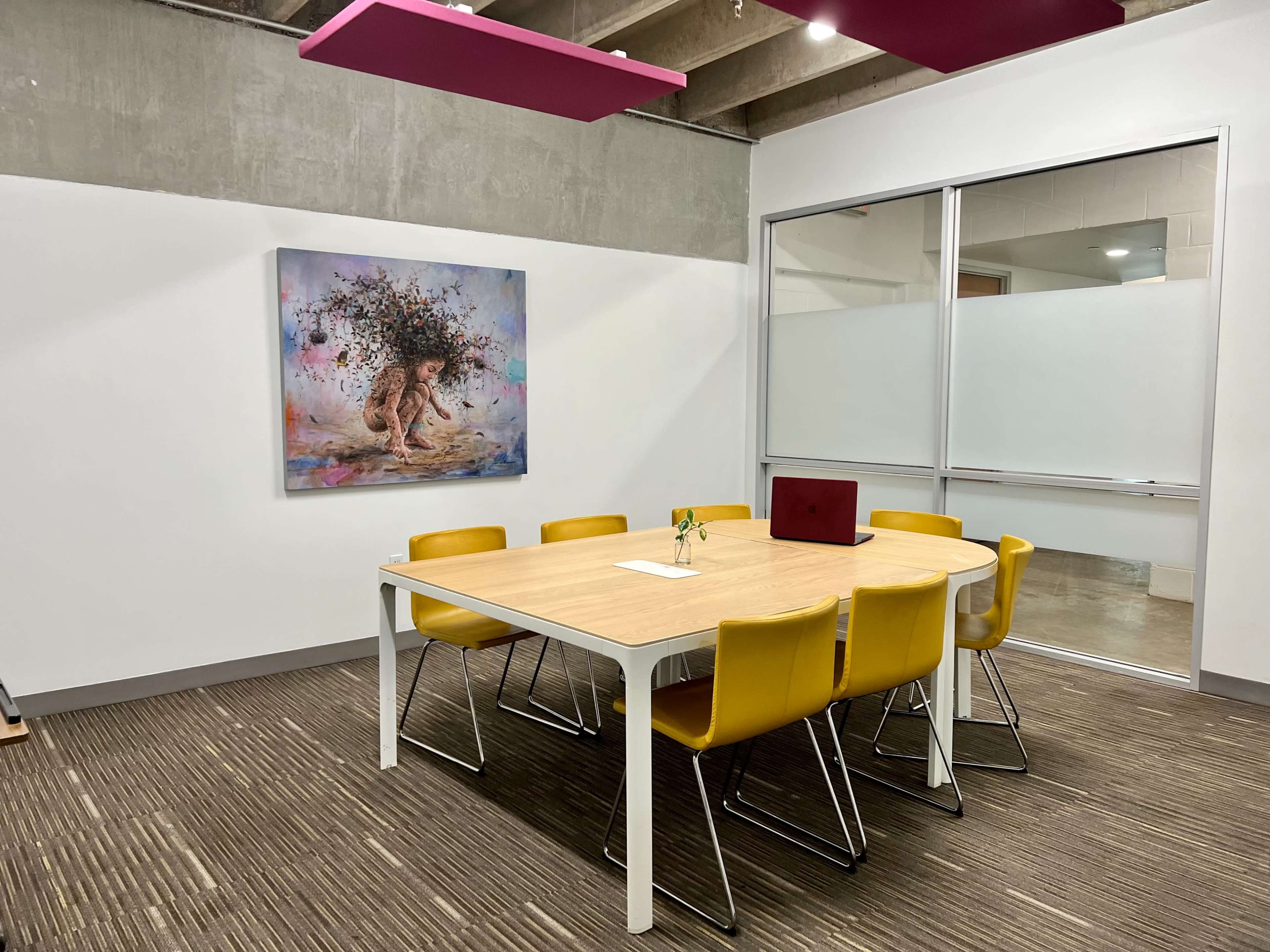 A modern conference room features a wooden table surrounded by six yellow chairs and a large artwork on the wall.