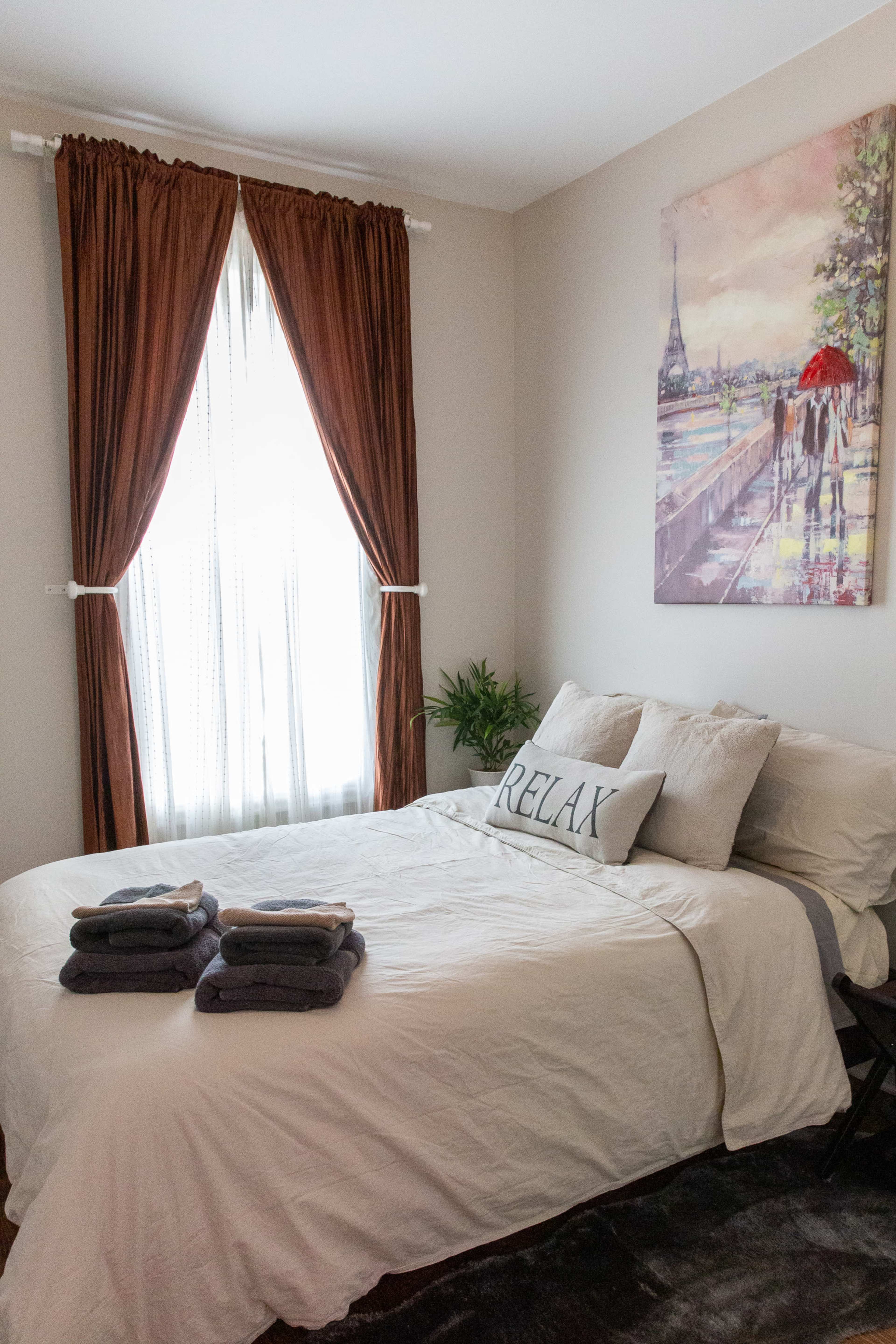 A neatly arranged bedroom features a bed with stacked towels, a decorative pillow that reads "RELAX," and a window draped with brown curtains.