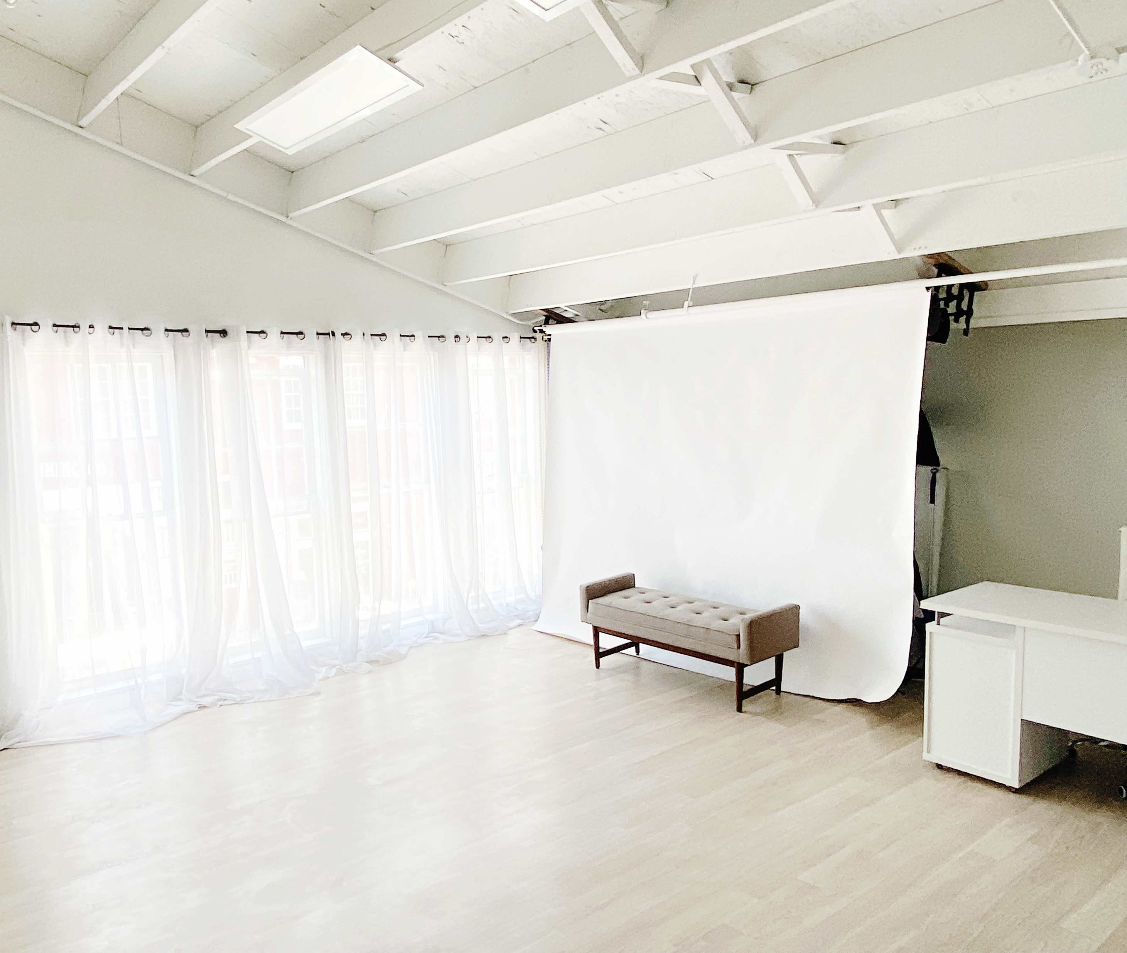 A bright, spacious room features large windows with sheer white curtains, a light-colored wooden floor, a simple beige bench, and a white backdrop against one wall.