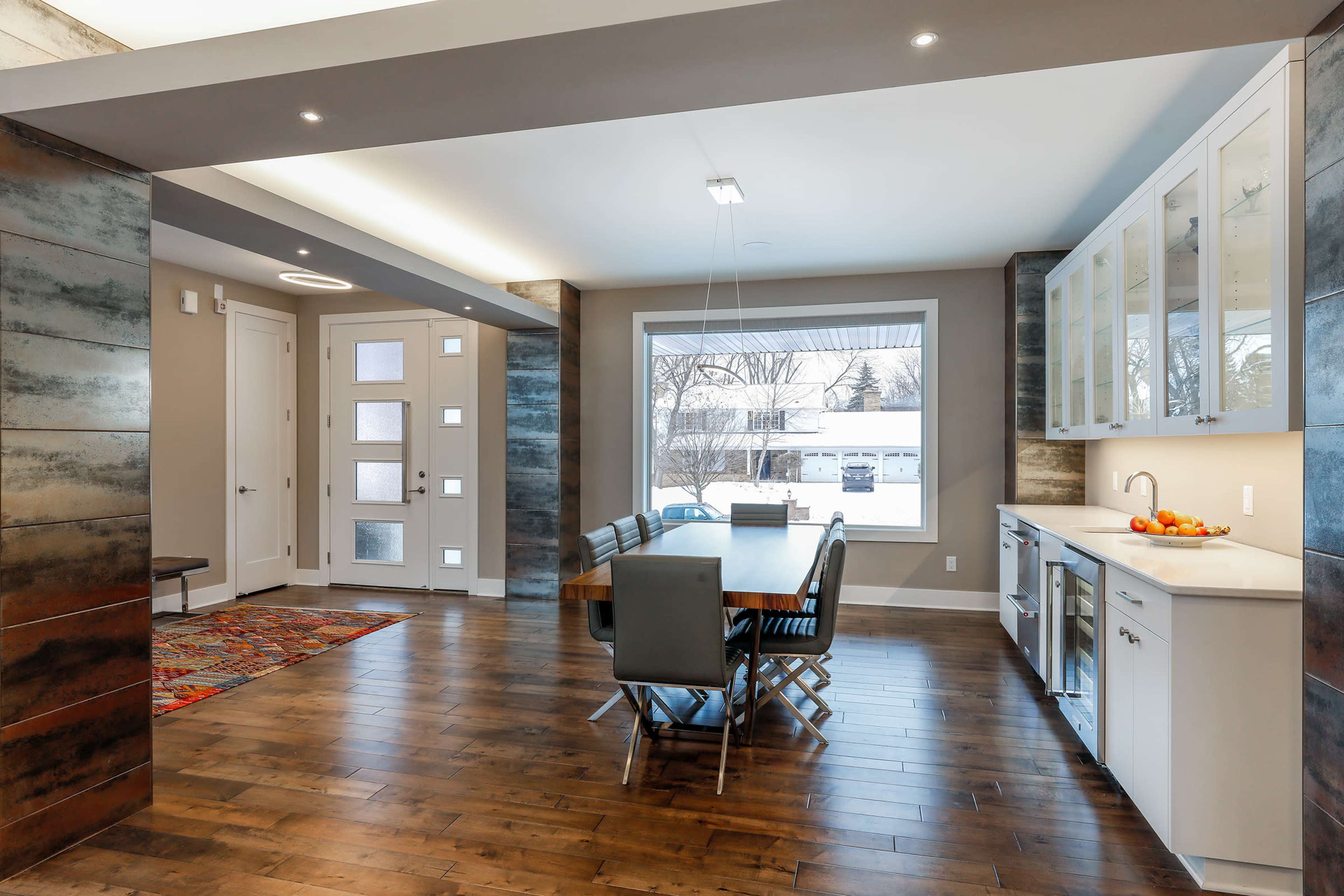 A modern dining area features a wooden table surrounded by black chairs, adjacent to a kitchen with white cabinets and a large window overlooking a snowy exterior.