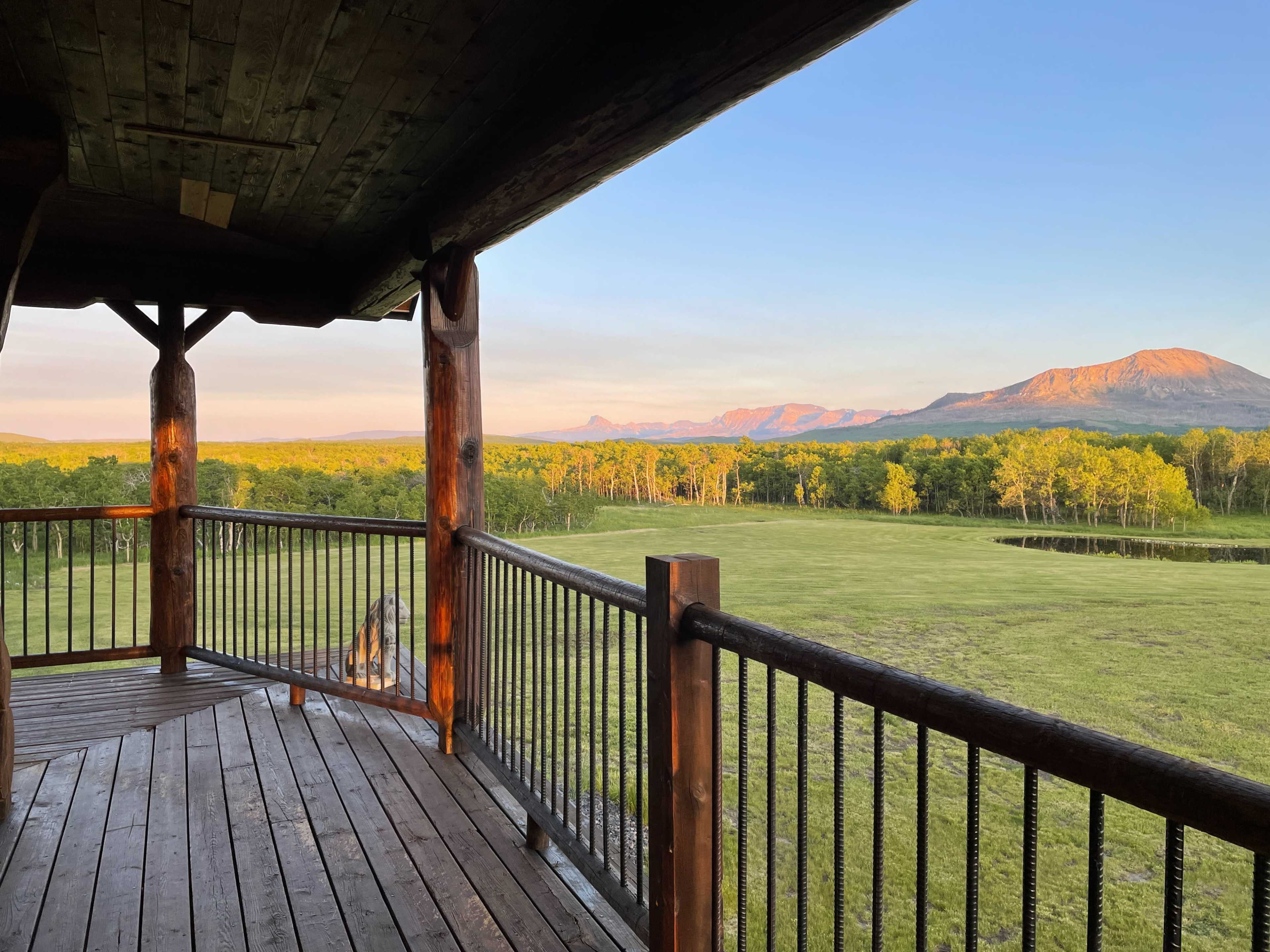 The image shows a wooden deck overlooking a grassy area and mountains under a clear sky during sunrise.