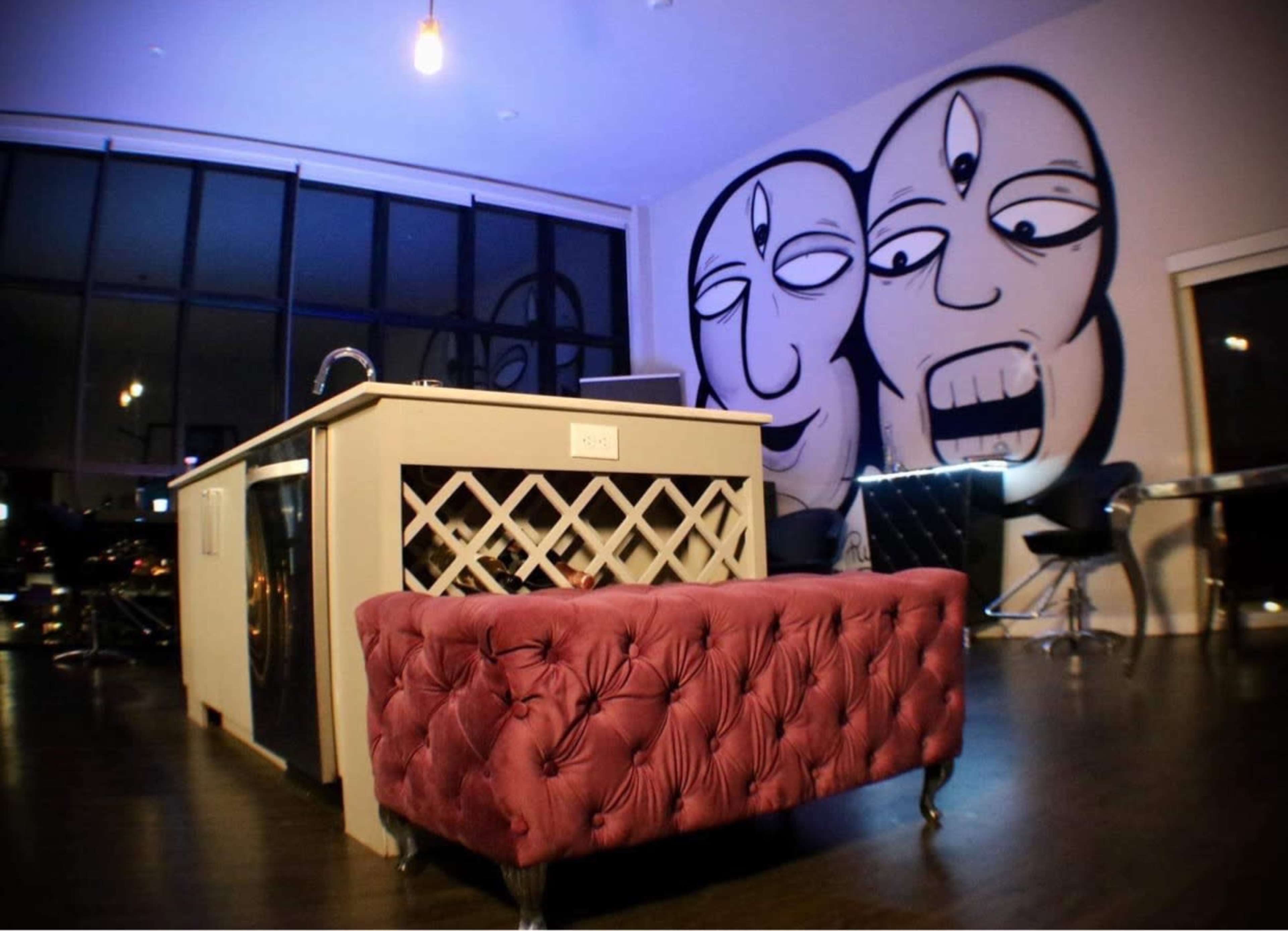 A modern interior featuring a plush burgundy bench and a large wall mural of two stylized faces, with a kitchen island and dining area visible in the background.