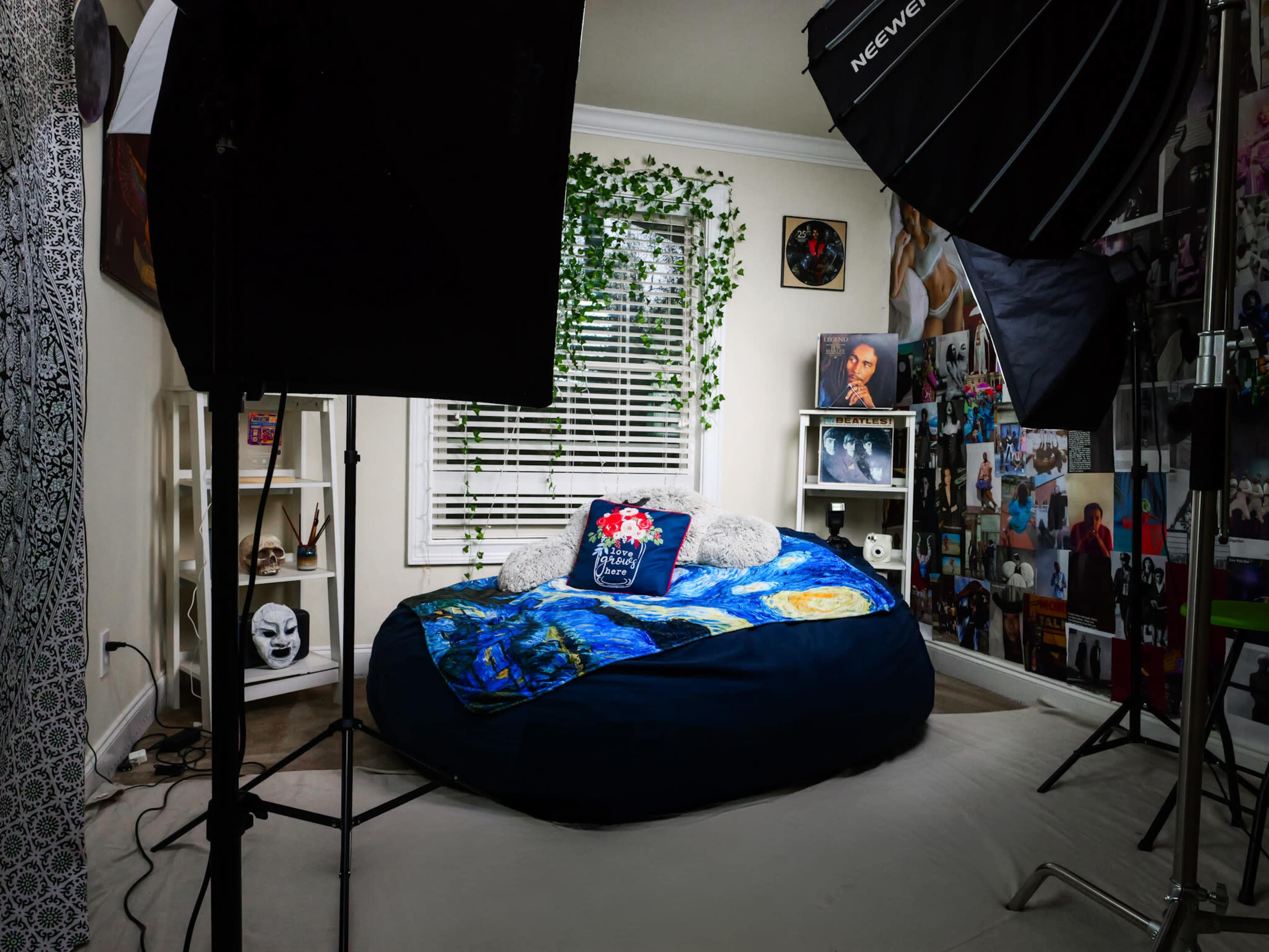 The image depicts a cozy bedroom with a large circular bed covered in a colorful blanket, surrounded by photography backdrops, lighting equipment, and decorative plants.