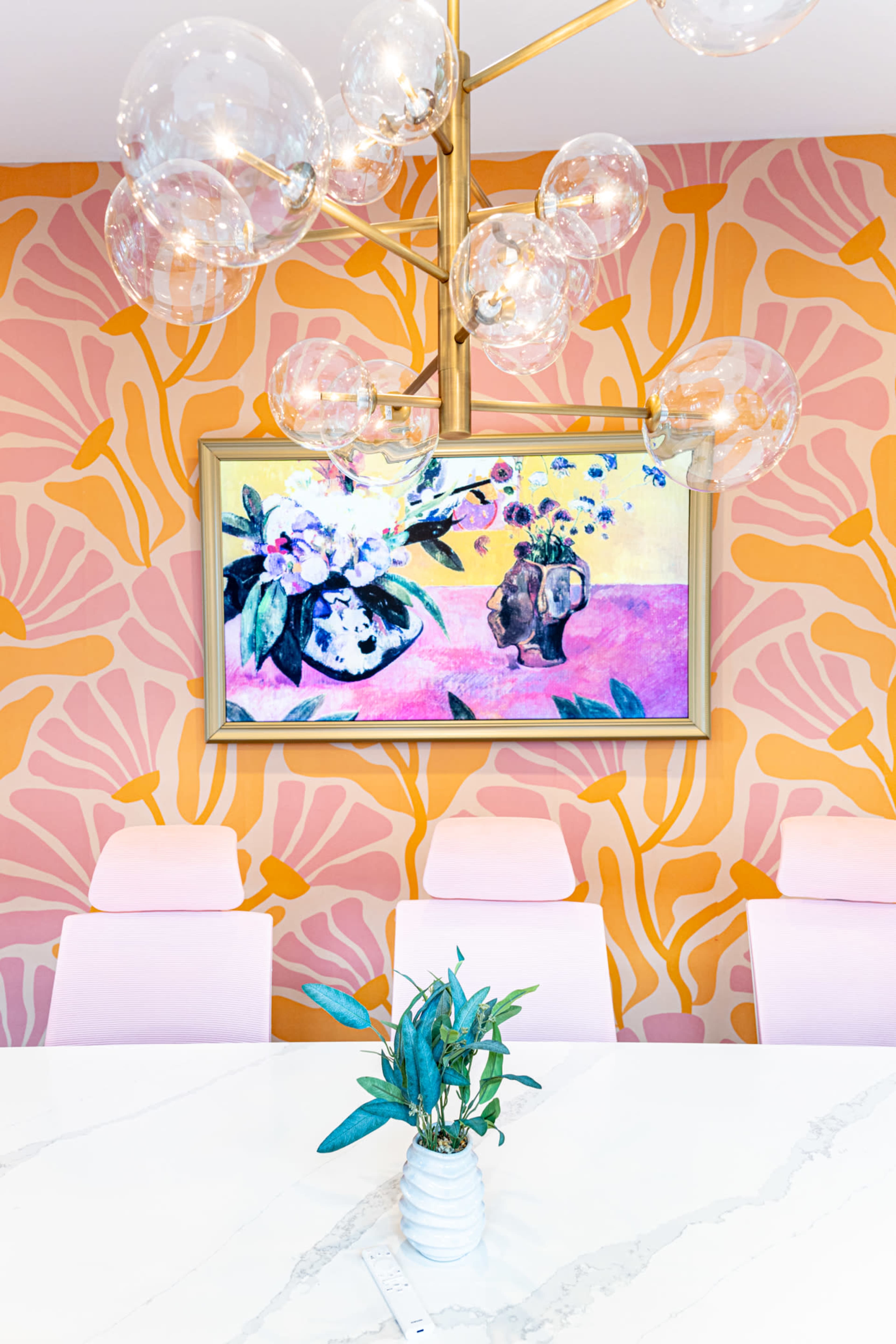 A dining area features a colorful floral wall mural, a modern chandelier with glass bulbs, and a table set with a small potted plant.