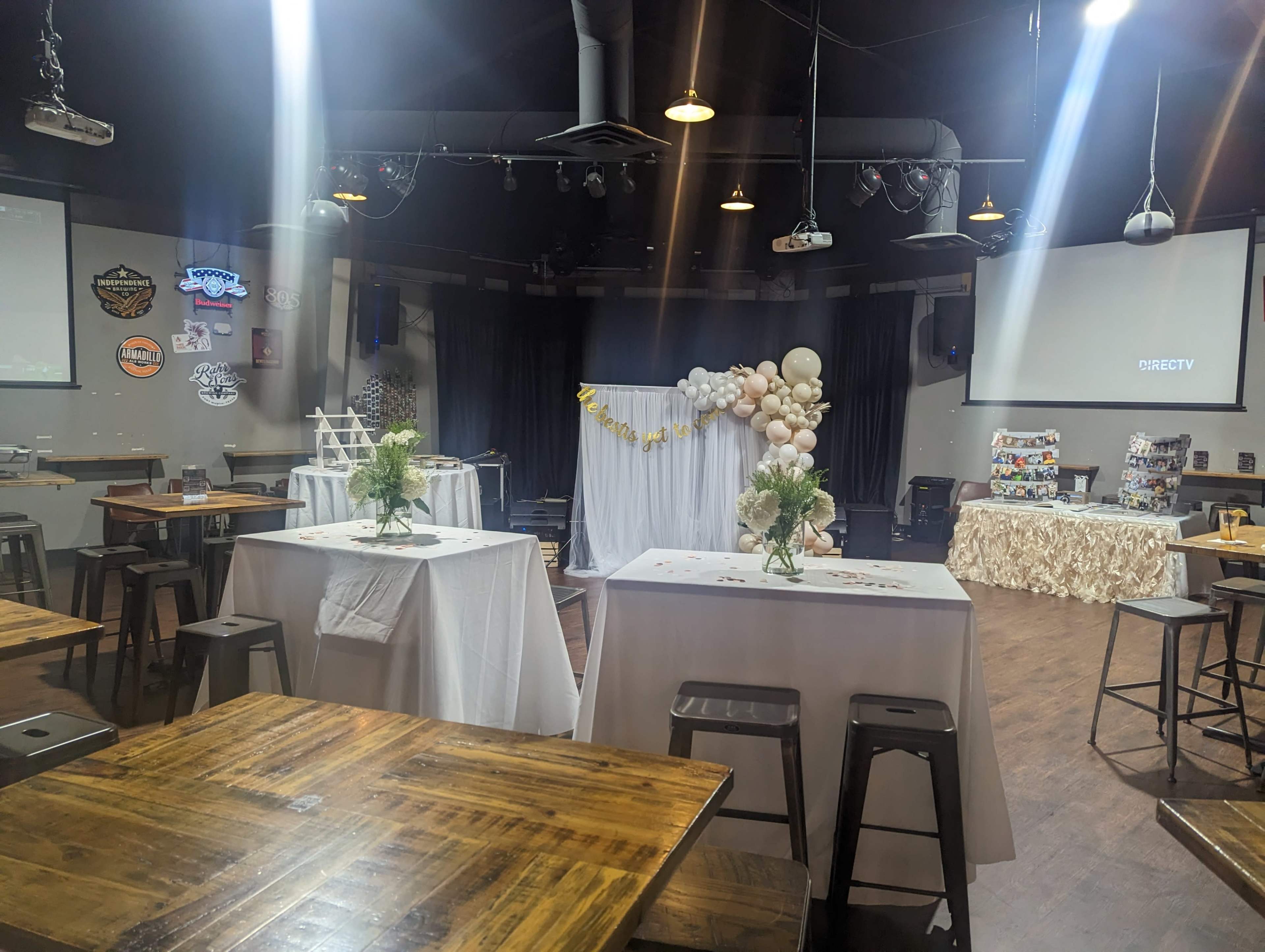 A spacious venue set up for an event, featuring tables with white tablecloths, floral centerpieces, a decorated backdrop, and screens displaying presentations.