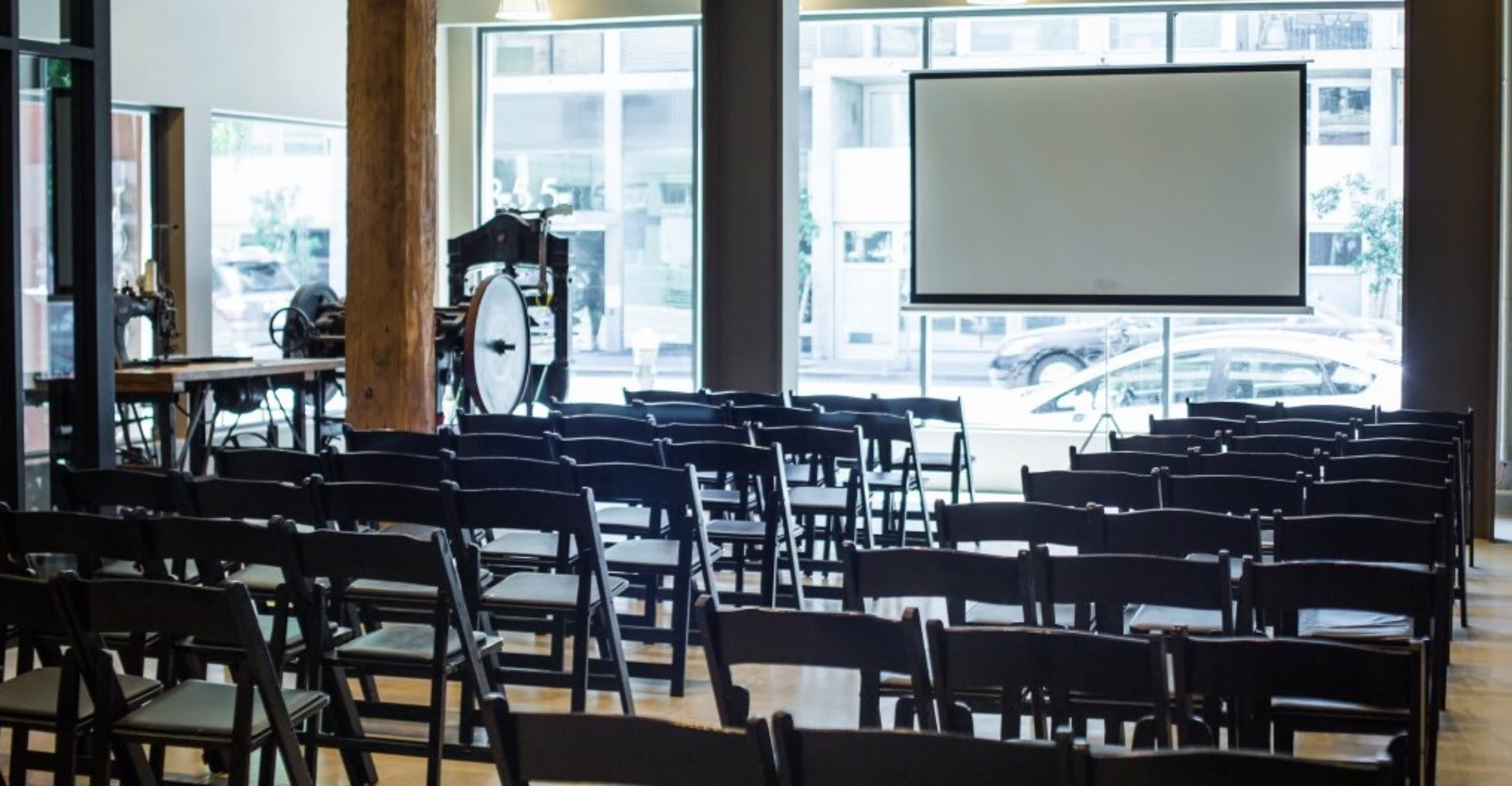 The image shows an empty auditorium with rows of black chairs facing a large screen, illuminated by natural light from large windows.
