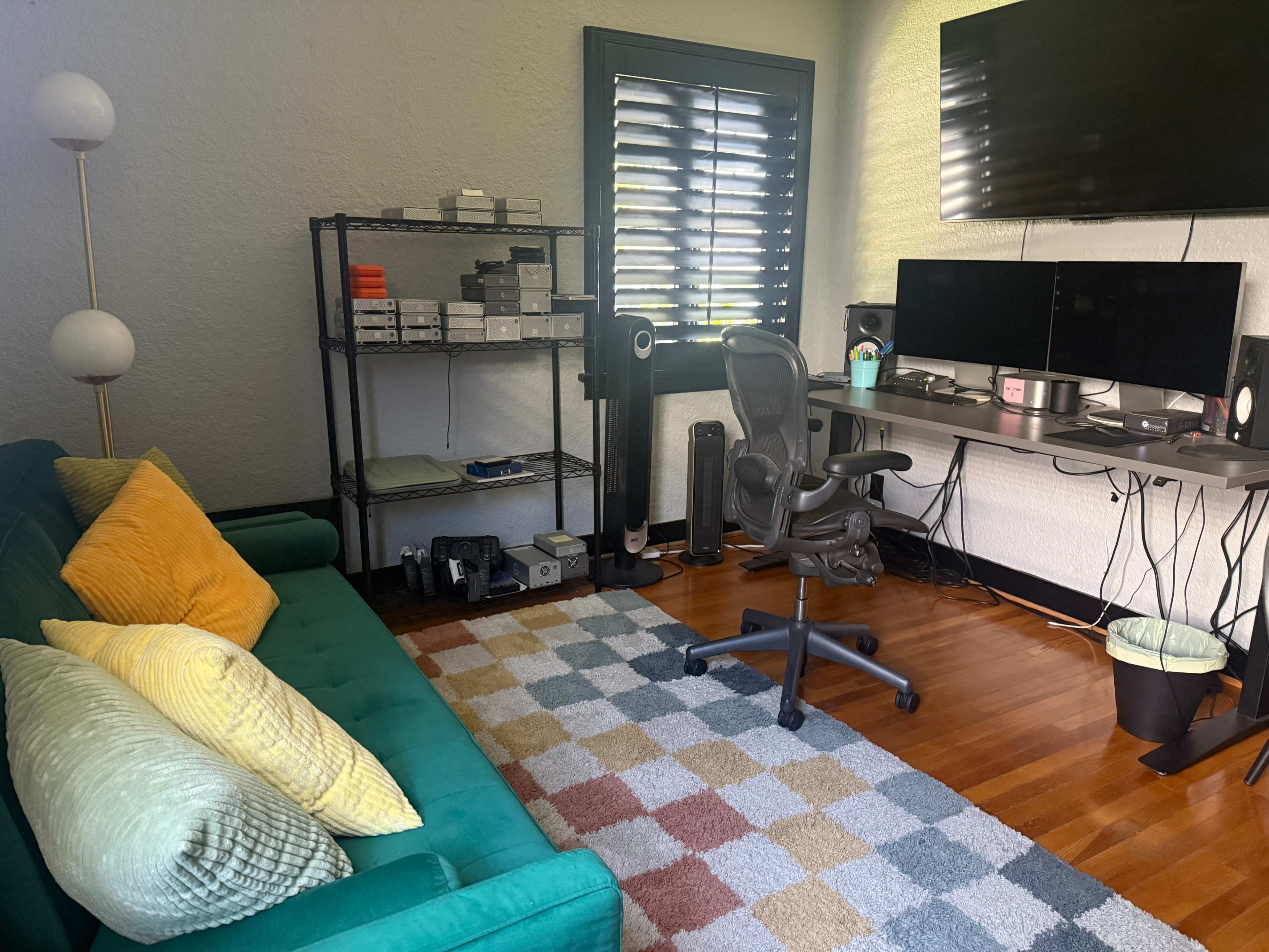 The image shows a neatly arranged home office with a green sofa, a desk featuring two monitors, and a shelf with various electronic devices and a floor lamp.