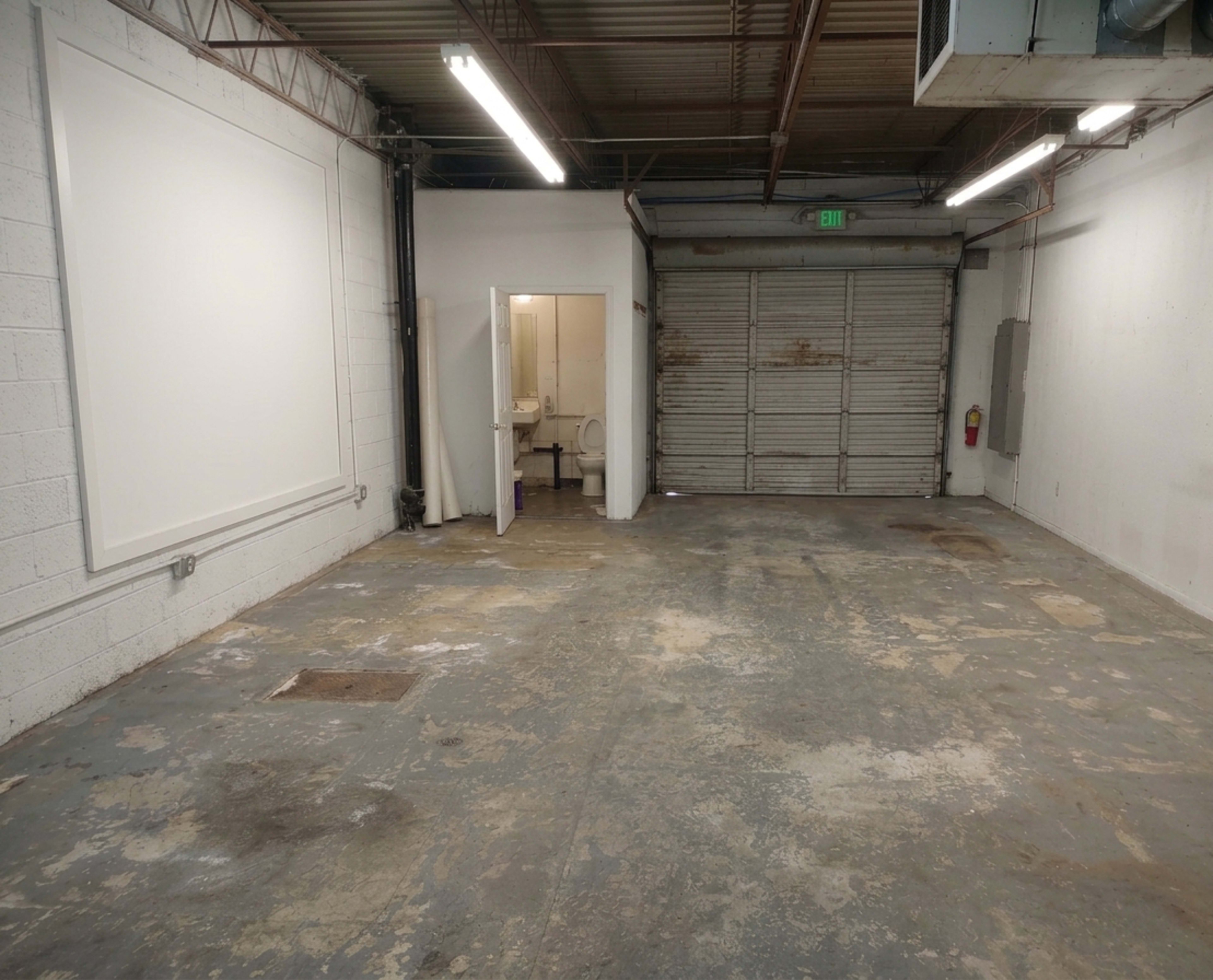 The image shows an empty industrial space with concrete flooring, a set of double doors on one side, and a small bathroom area at the rear.
