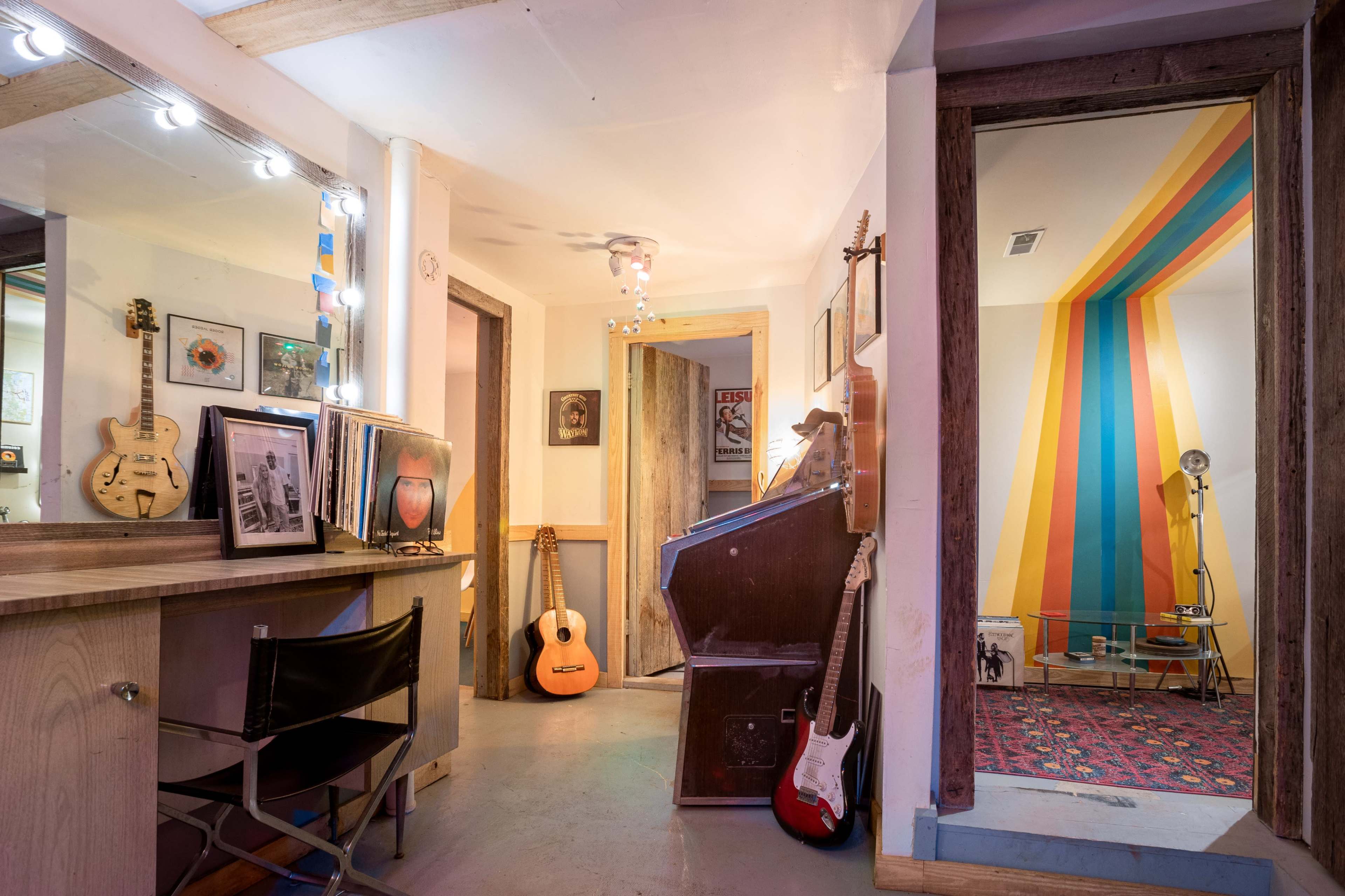 A well-lit interior space featuring a wooden desk, guitars, framed artwork, and a colorful striped wall in an adjacent room.