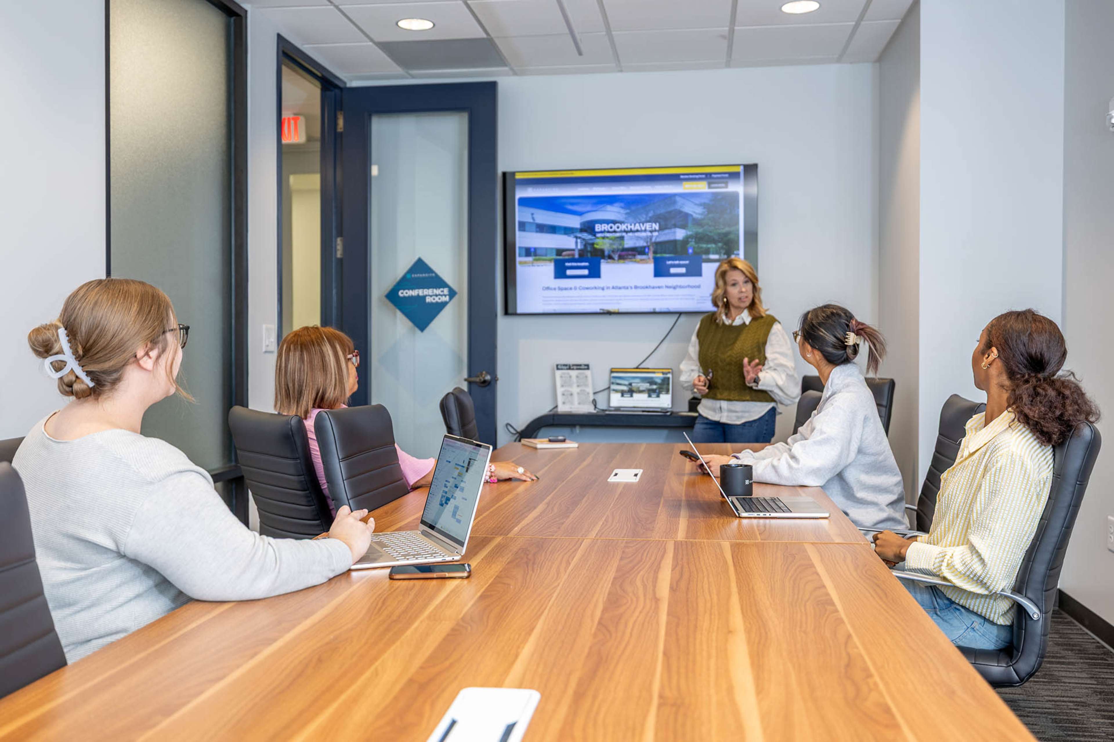 A group of five people is seated around a conference table, with one person standing and giving a presentation using a screen display.