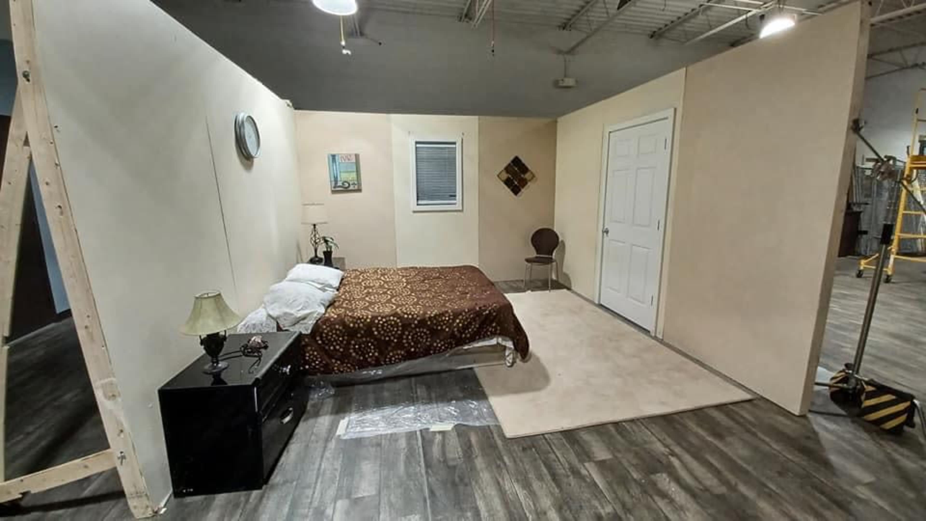 The image shows a sparsely furnished room with a bed, nightstand, and a chair, surrounded by plain walls and a window.