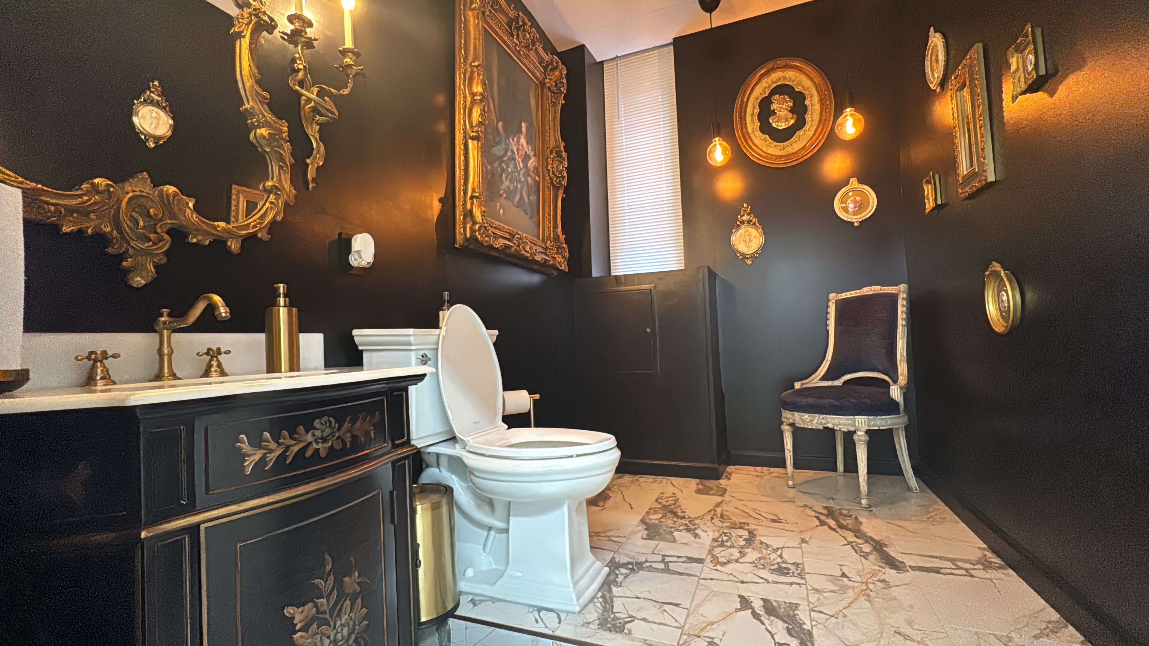 The image shows a darkly themed bathroom featuring a white toilet, a vintage-style chair, a marble floor, and several framed wall decorations.