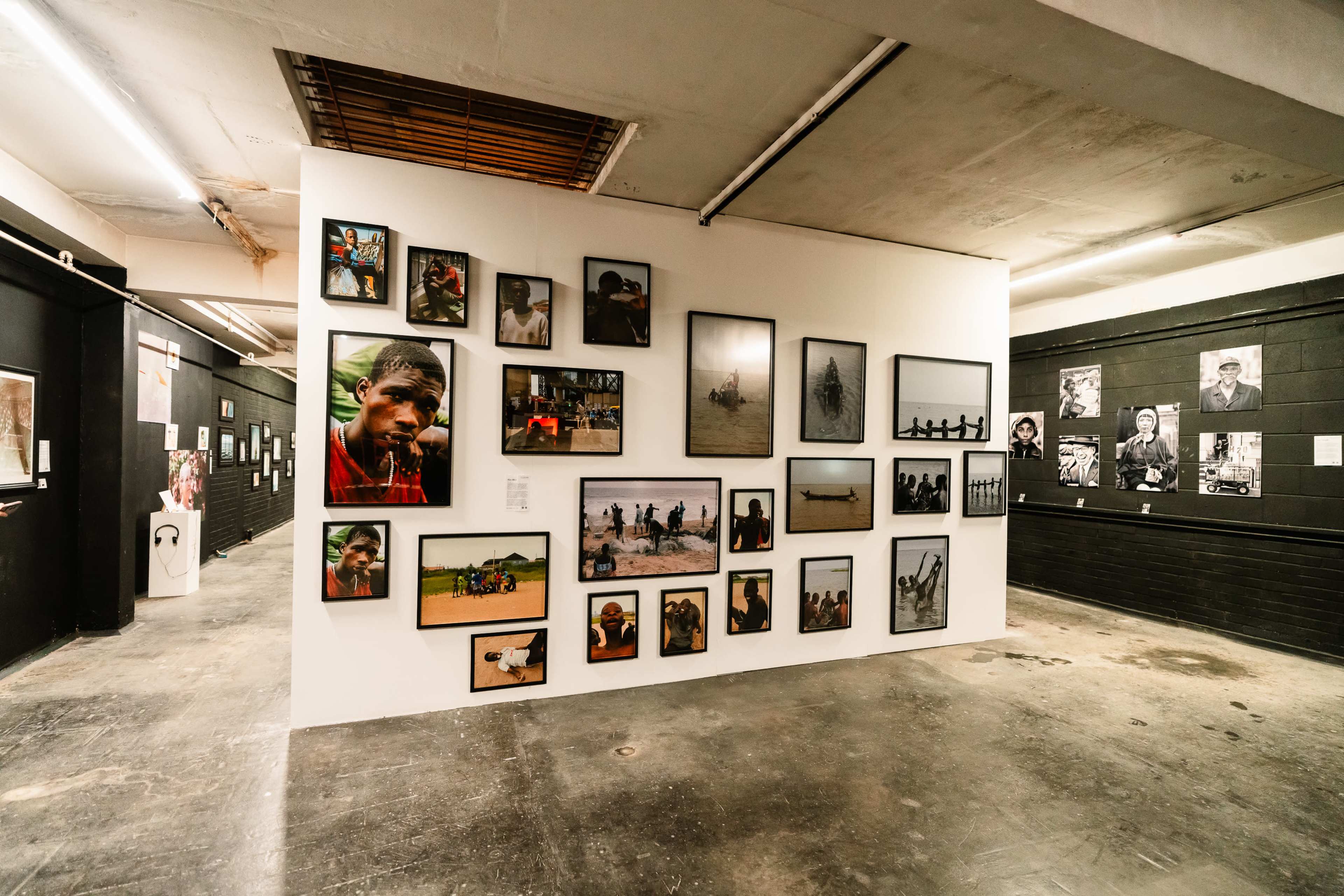 The image shows an art gallery with a white wall displaying various framed photographs, surrounded by darker walls with additional photographs and artwork.