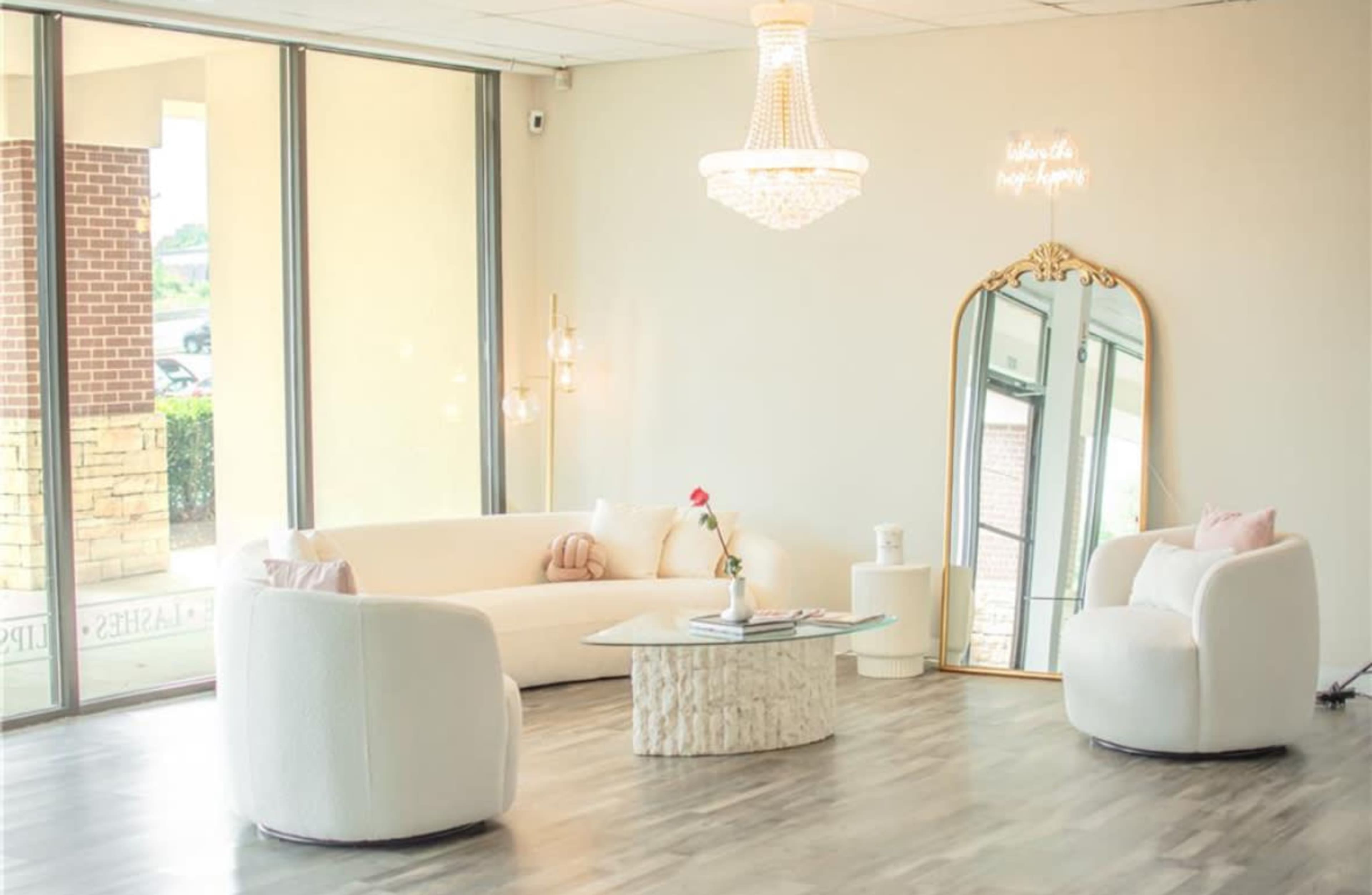 A modern seating area features white sofas, a glass coffee table, and a large mirror, illuminated by a chandelier and surrounded by large windows.